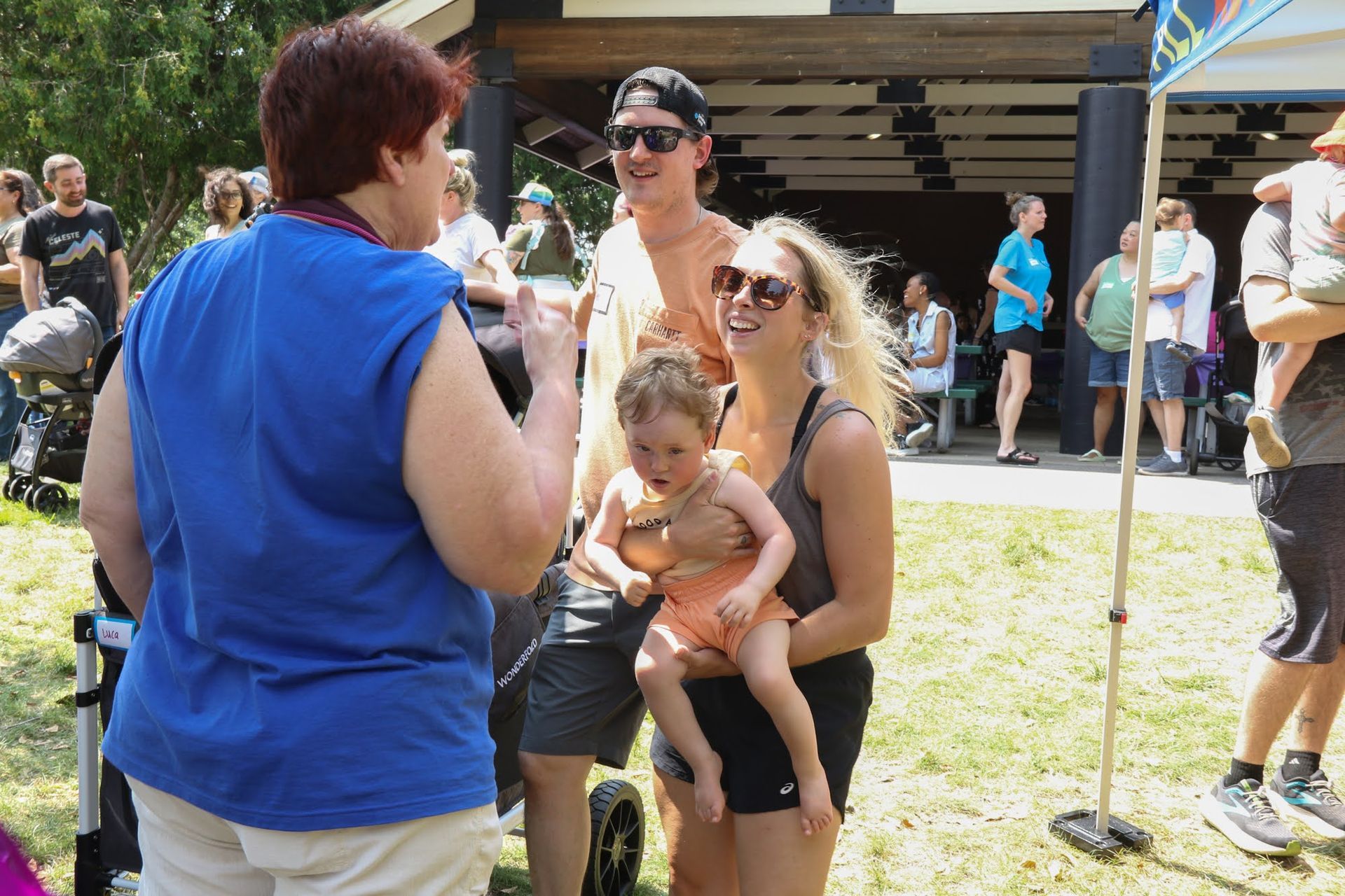 People talking outdoors, woman in blue shirt, woman holding baby, man wearing sunglasses, sunny day.