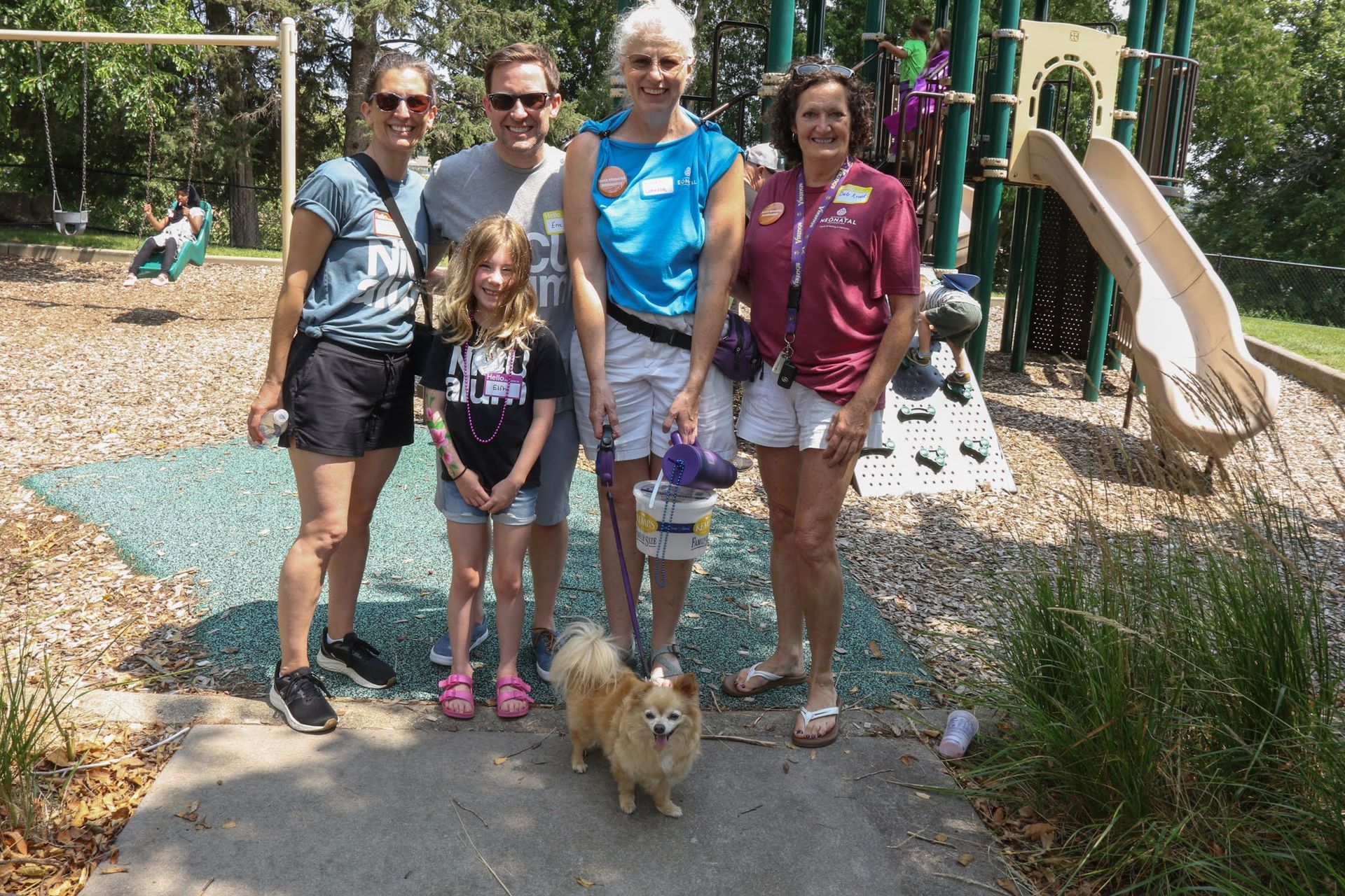 Group of people and a dog at a playground: adults, child, small dog, smiling, sunny day.