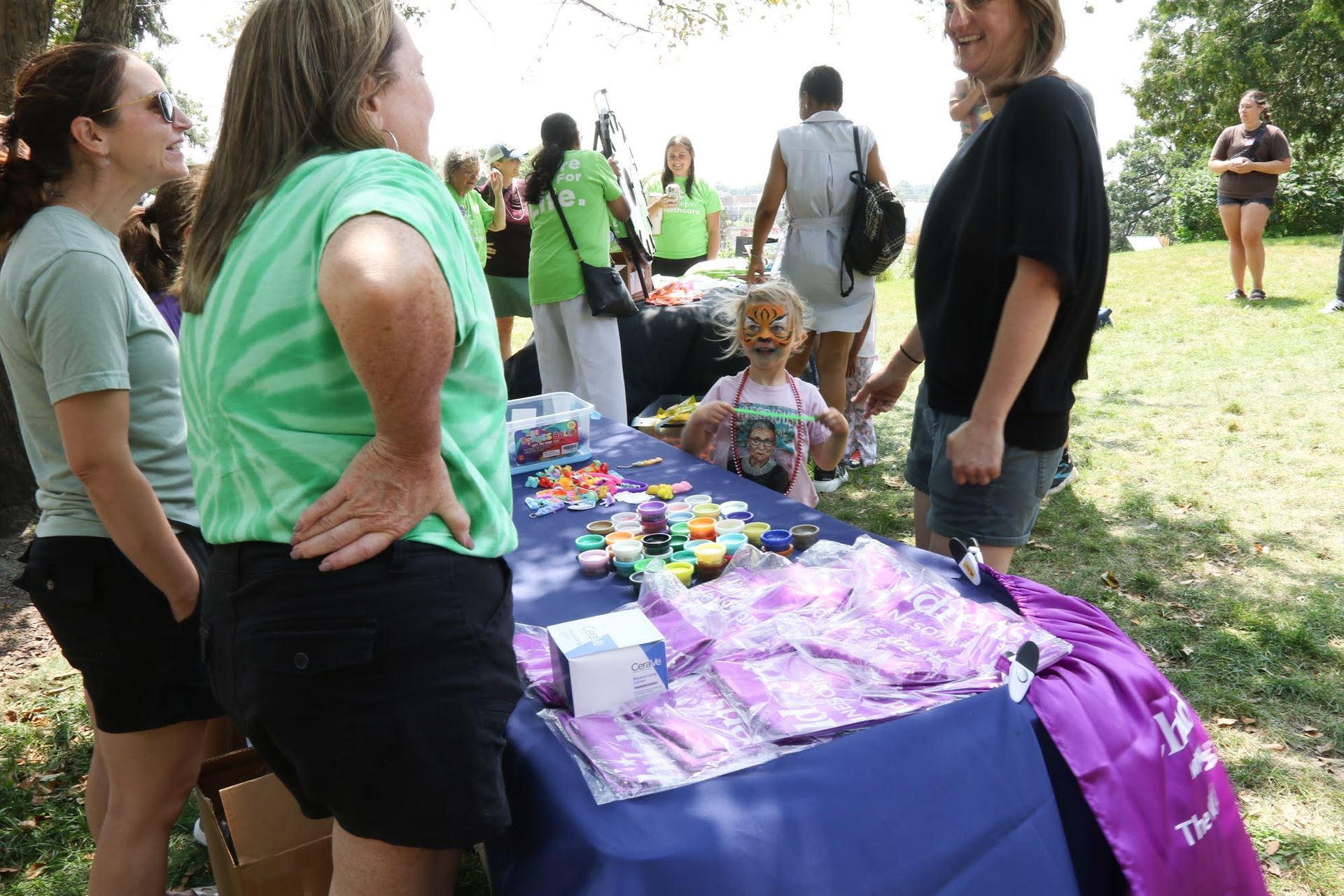 People at a table with art supplies outdoors. Child with face paint smiles. Others in shirts.