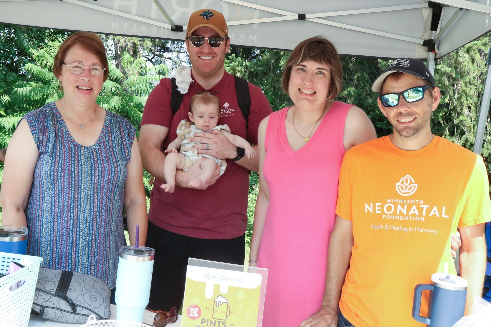Five people under a tent; one man holds a baby.  They stand behind a table with items, smiles, and sunny weather.