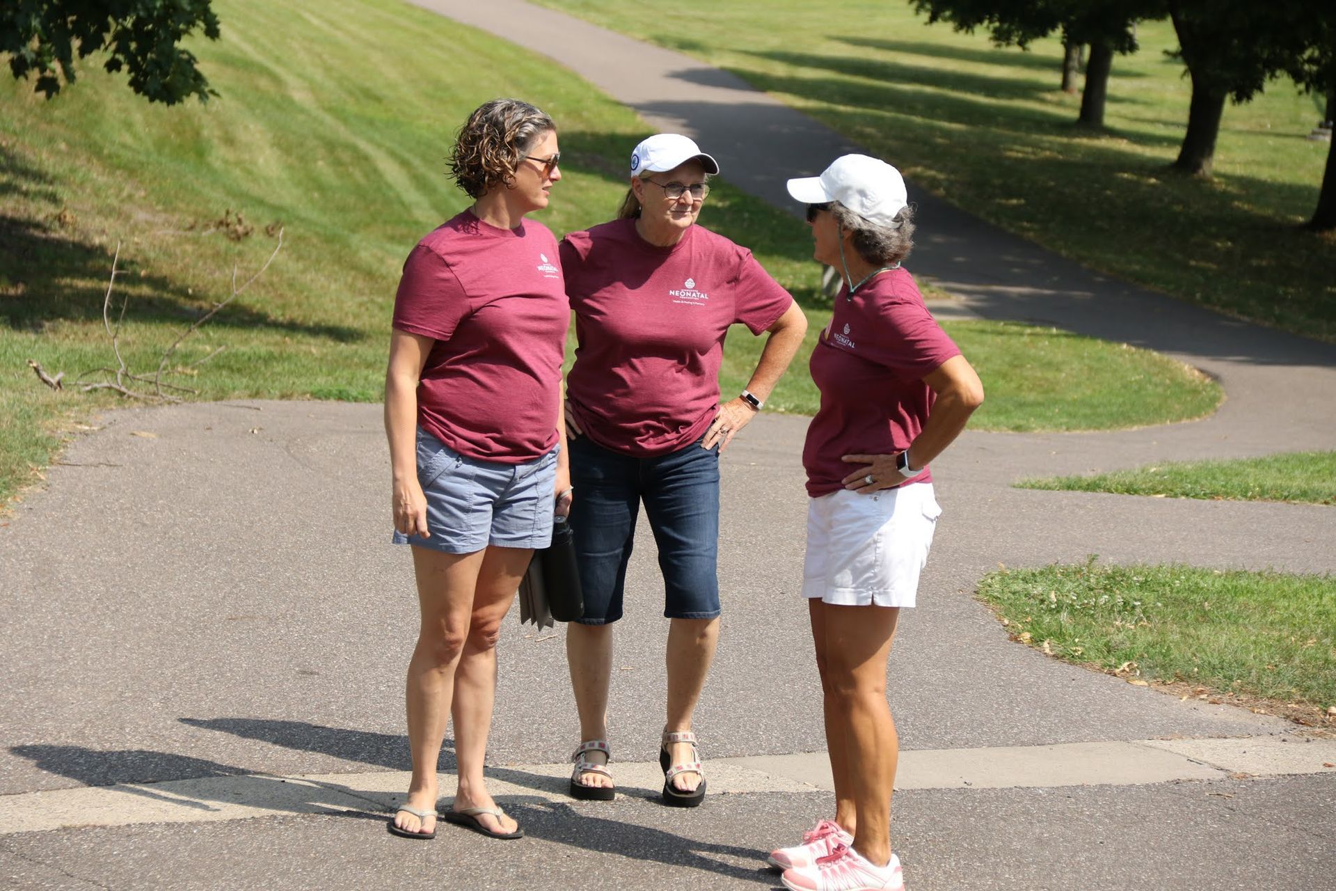 Three women in maroon shirts and hats stand on a paved path, talking.