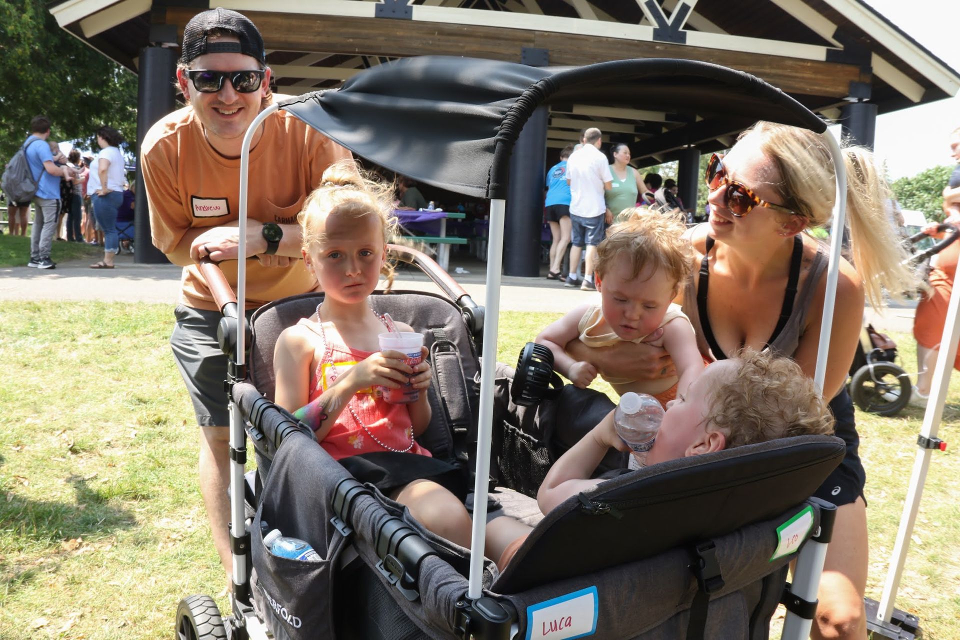 Family with three children in a wagon at an outdoor event, people and pavilion in background.