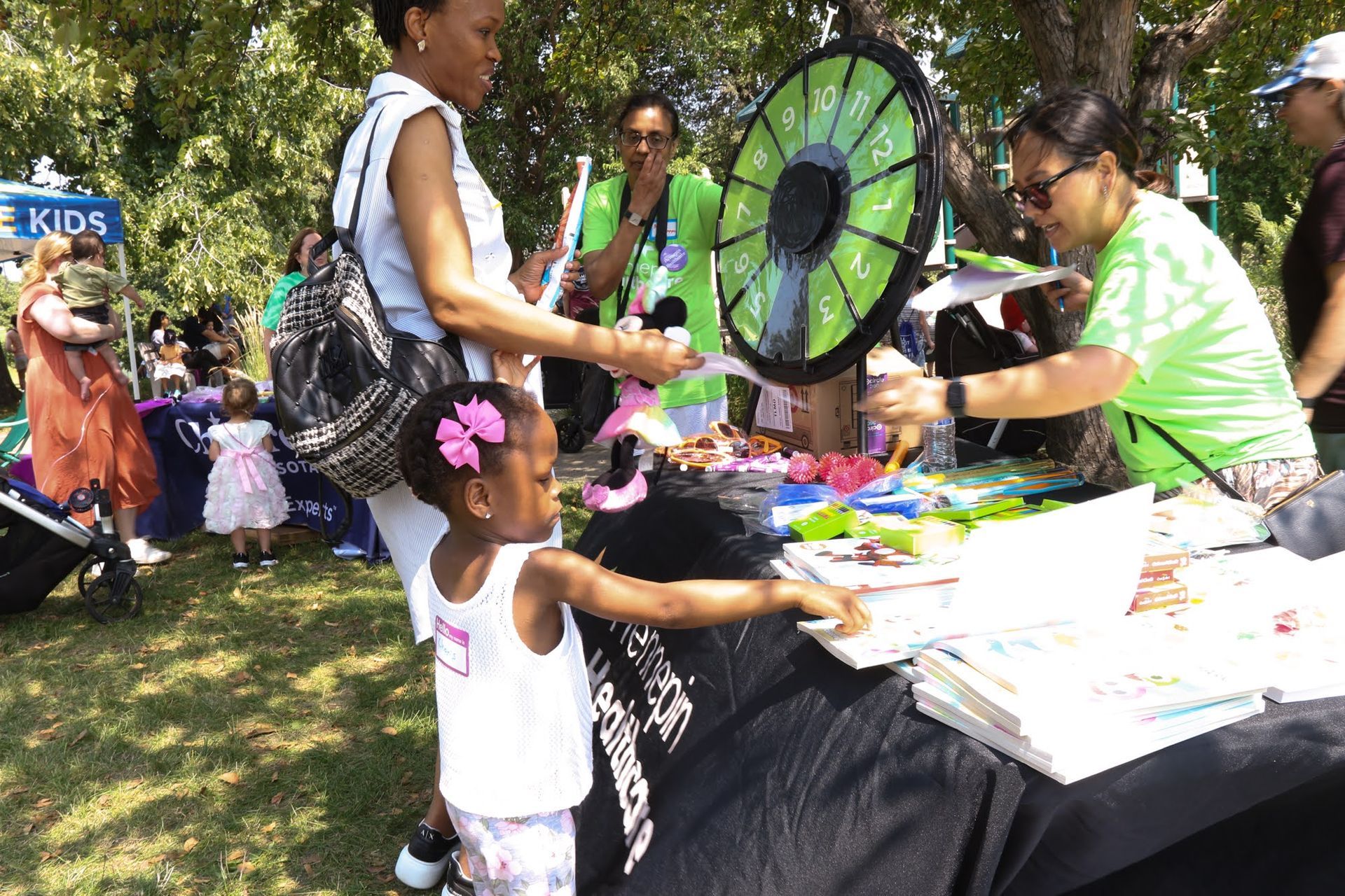 A child points to a prize at a fair booth with a prize wheel; woman in green shirt helps.