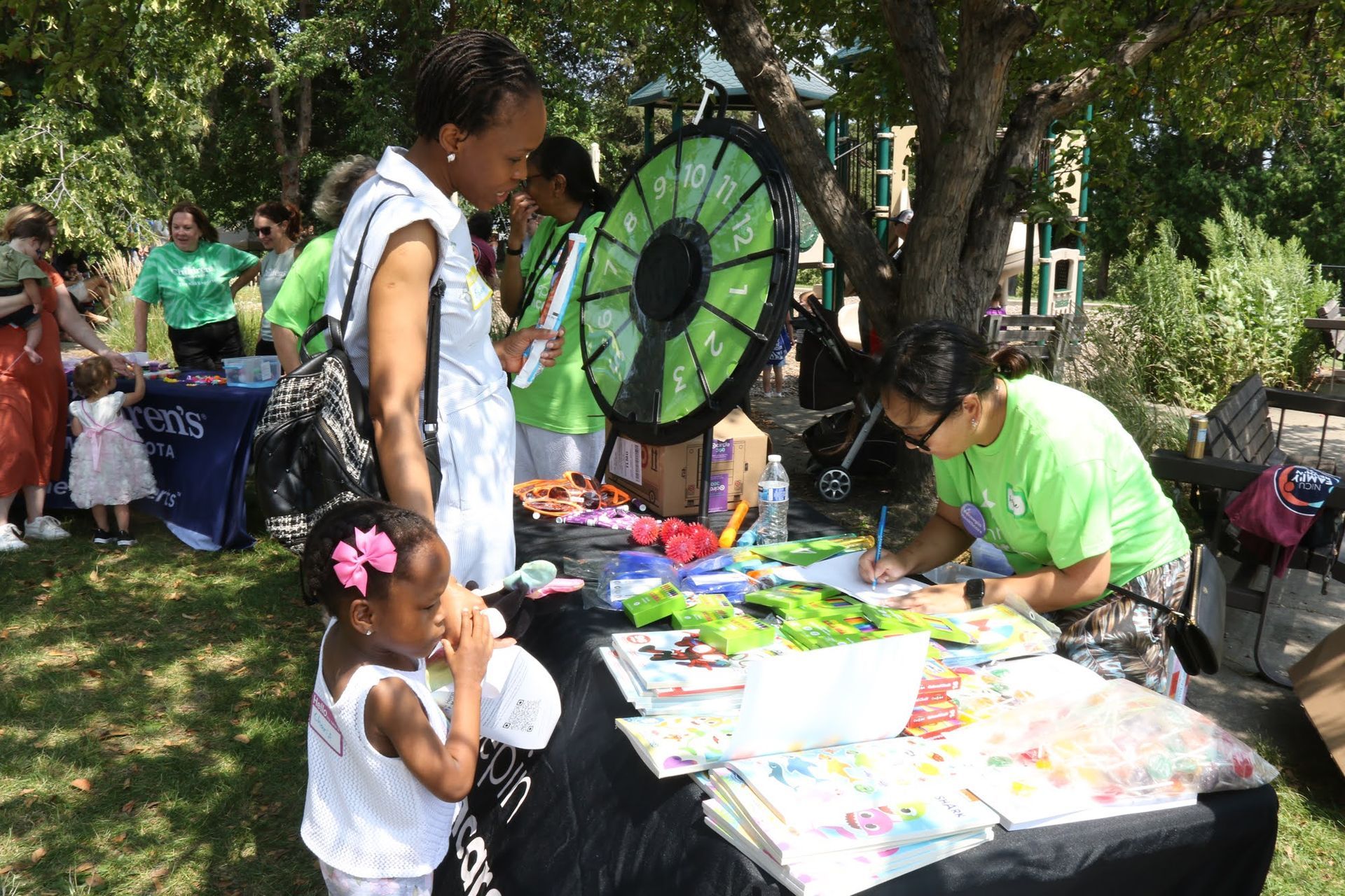 People at an outdoor event with a table of crafts and a prize wheel. A child eats a snack.
