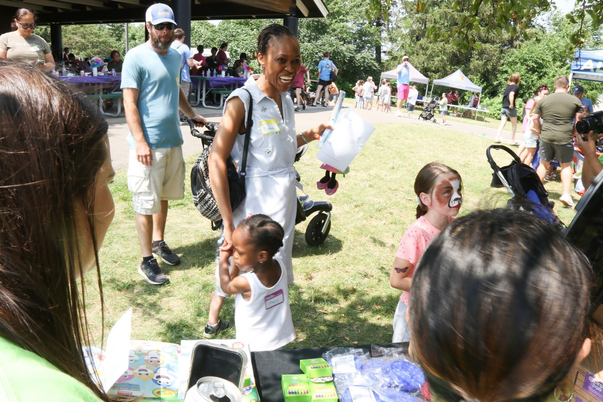 People at an outdoor event; a woman holds a container, children with face paint, a man with a stroller.