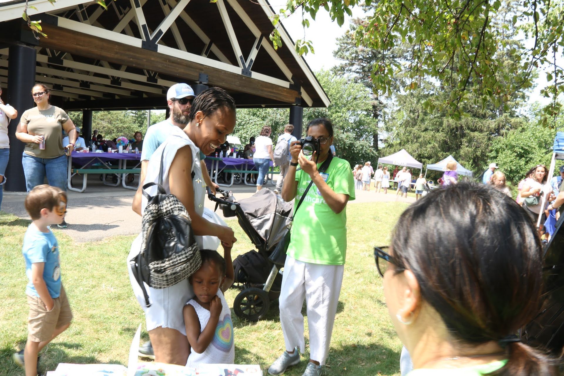 People at a park event. A woman poses with children as a man takes a photo. A stroller and pavilion are also present.