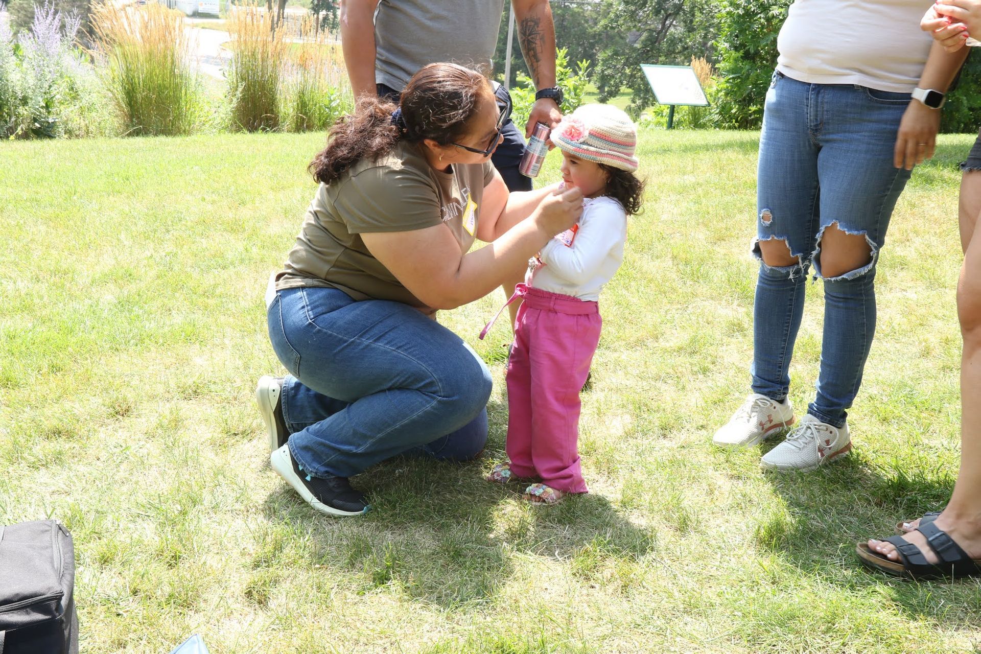 Woman kneeling, adjusting a small child's shirt on a grassy field; others standing nearby.