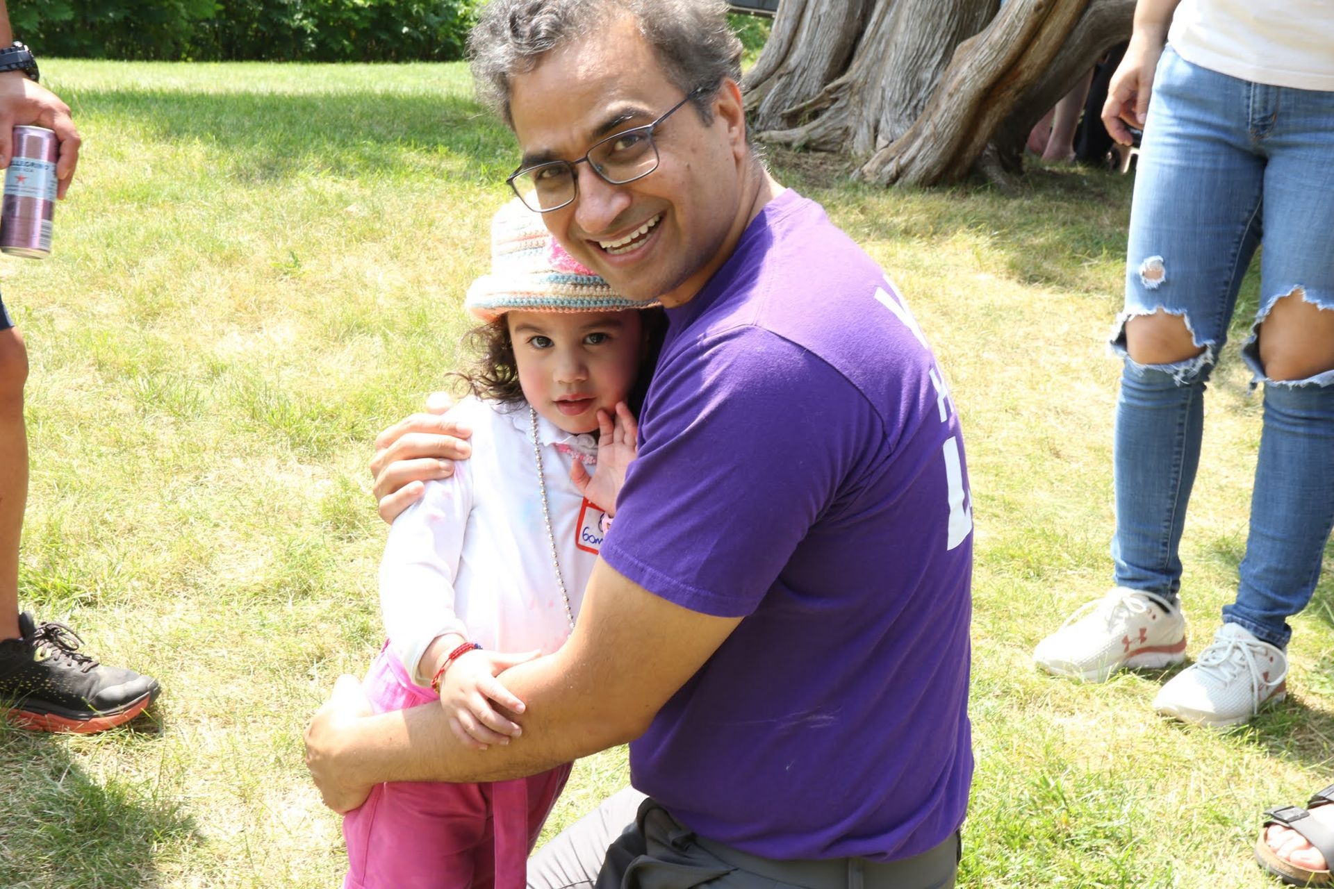 Man hugging a child outdoors; both smiling. The man wears purple, the child pink; sunny day.