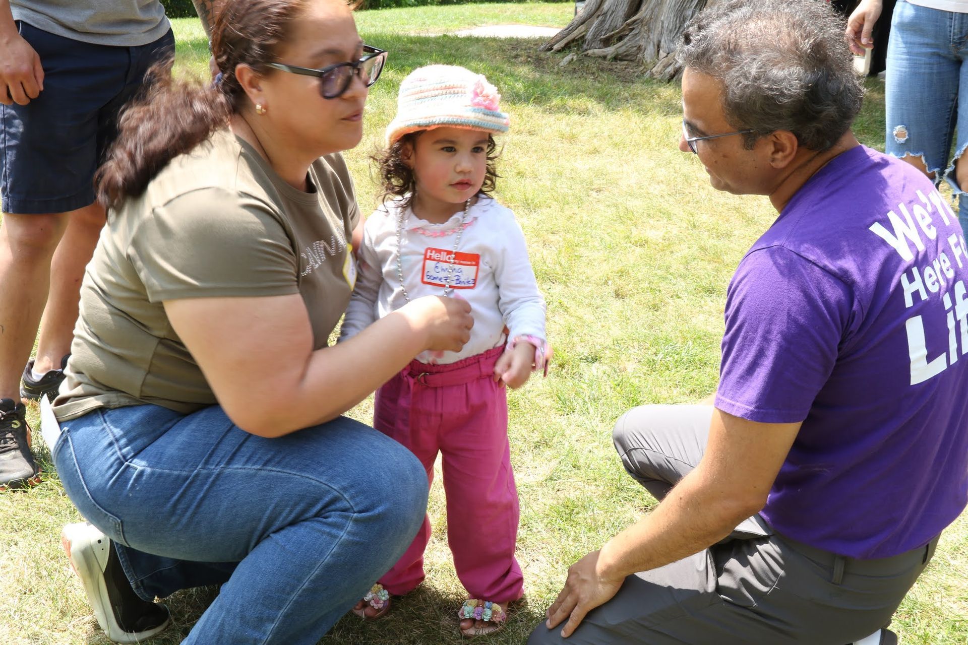 Woman and man kneeling, talking to a young child outdoors. Child wears a hat and pink pants.