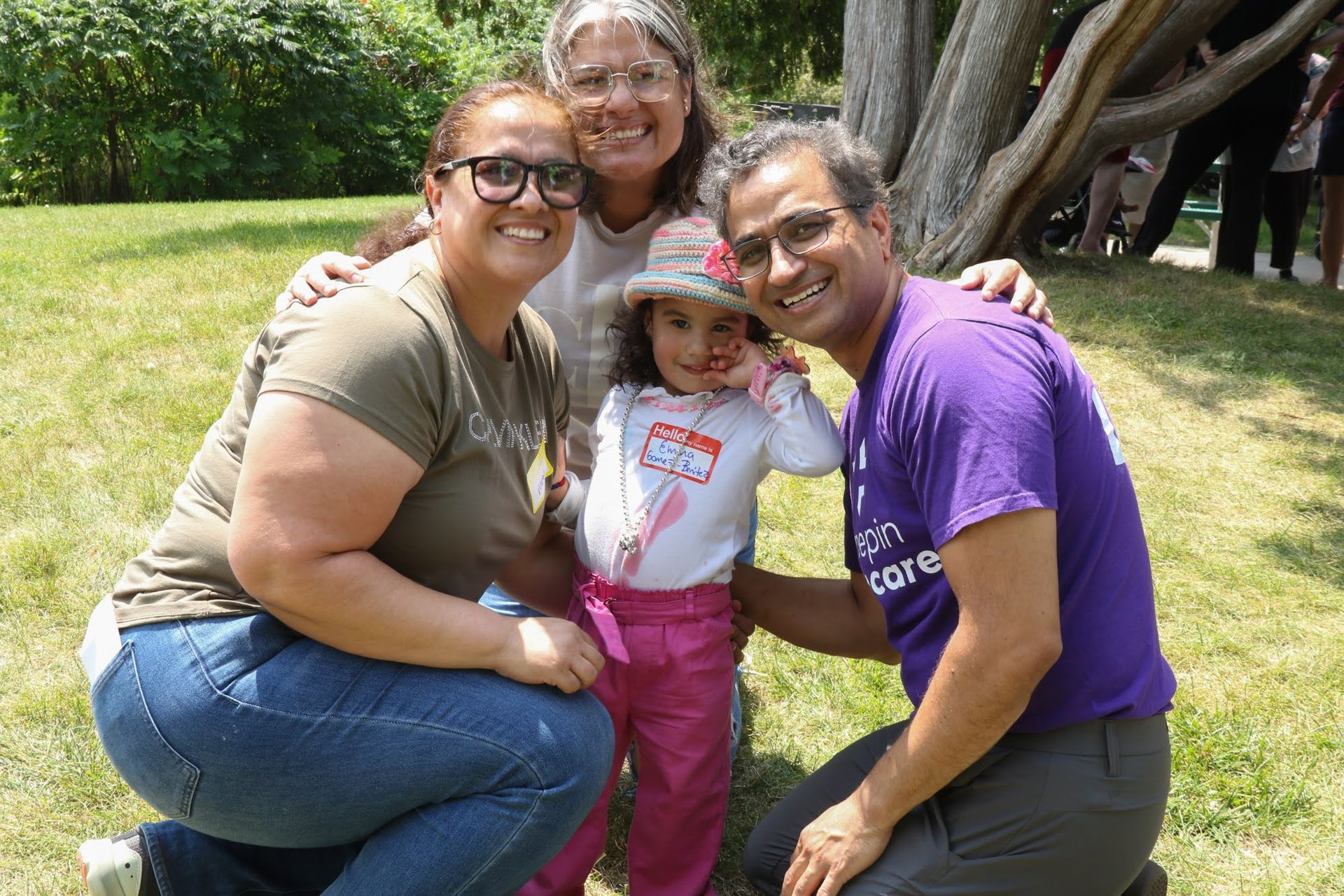 Four people smiling, posing for photo on grass, under tree. Girl in pink pants and shirt, tag.