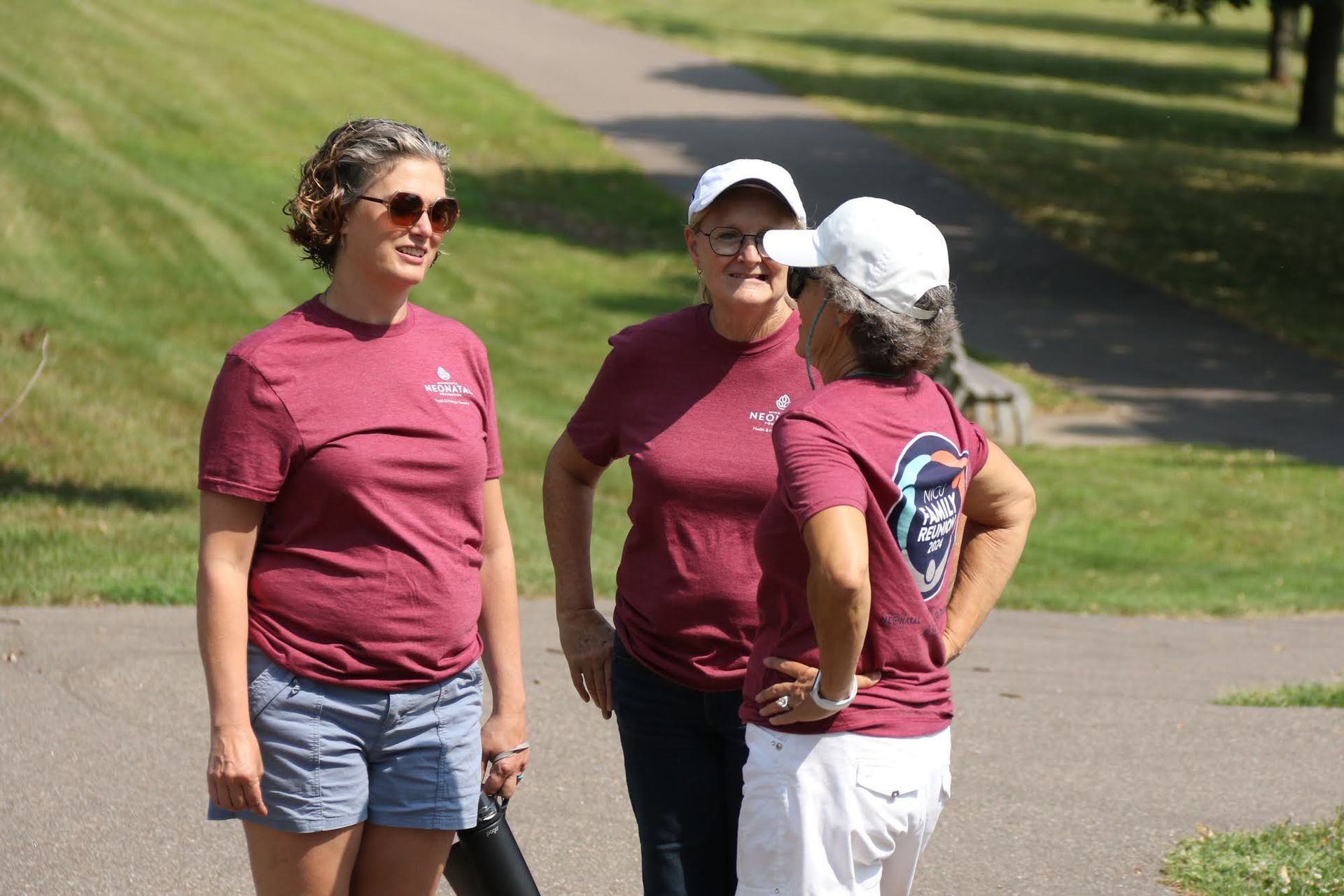 Three women in matching maroon shirts and hats talking on a paved path in a park.
