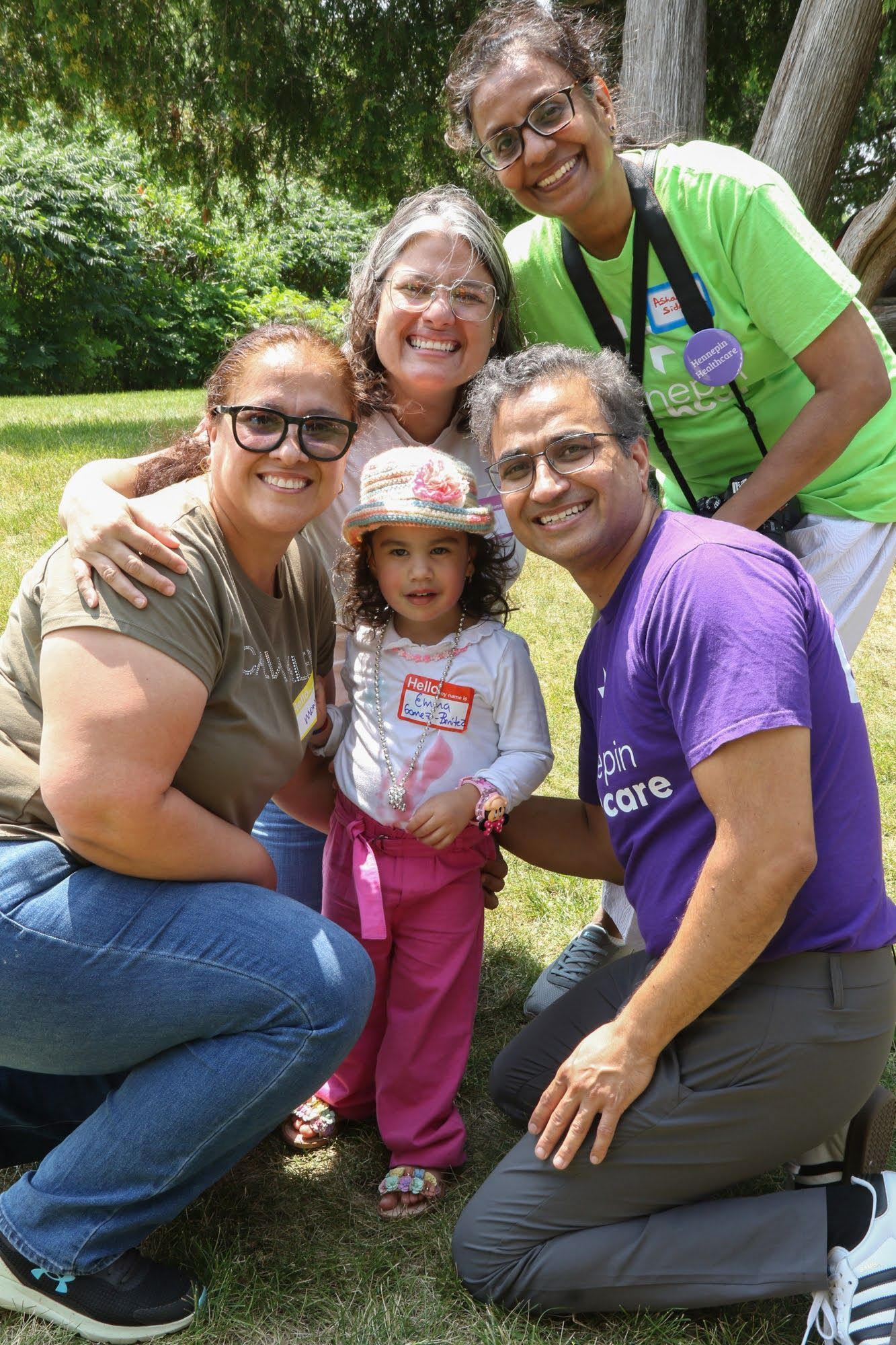 Group of five people smiling outdoors, surrounding a young child wearing a hat and pink pants.