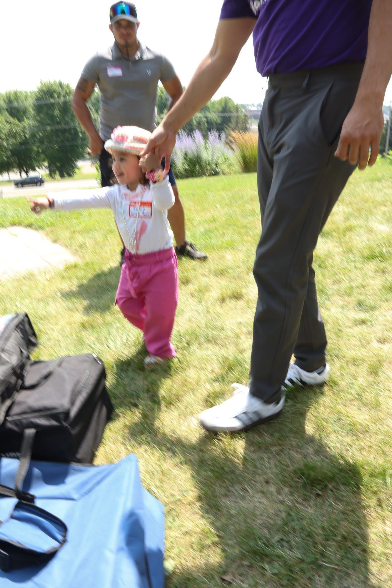Child with pink pants and hat points, man holds her hand, another man watches on grass.