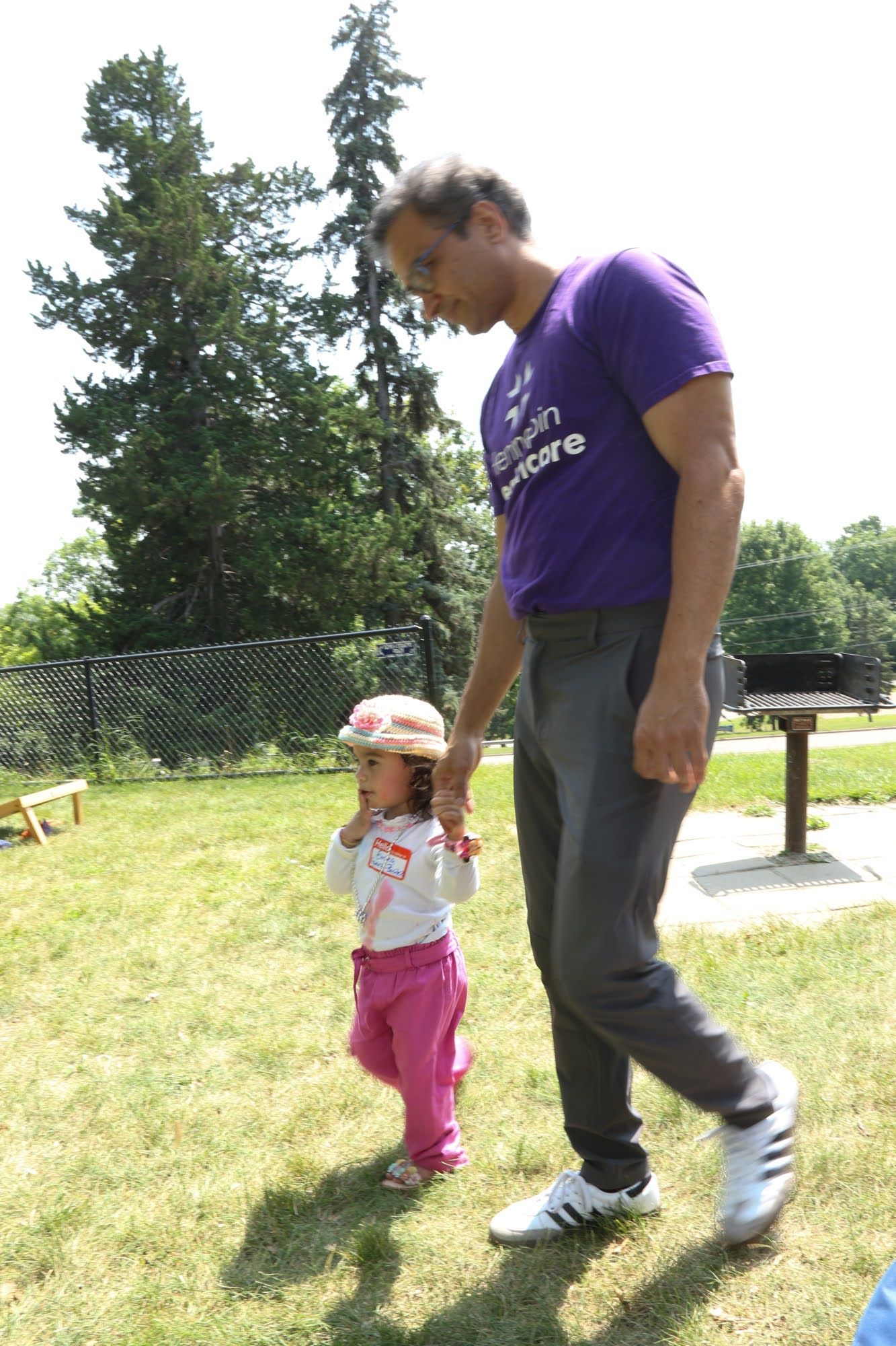 Man in purple shirt holds the hand of a toddler walking on grass in a park.