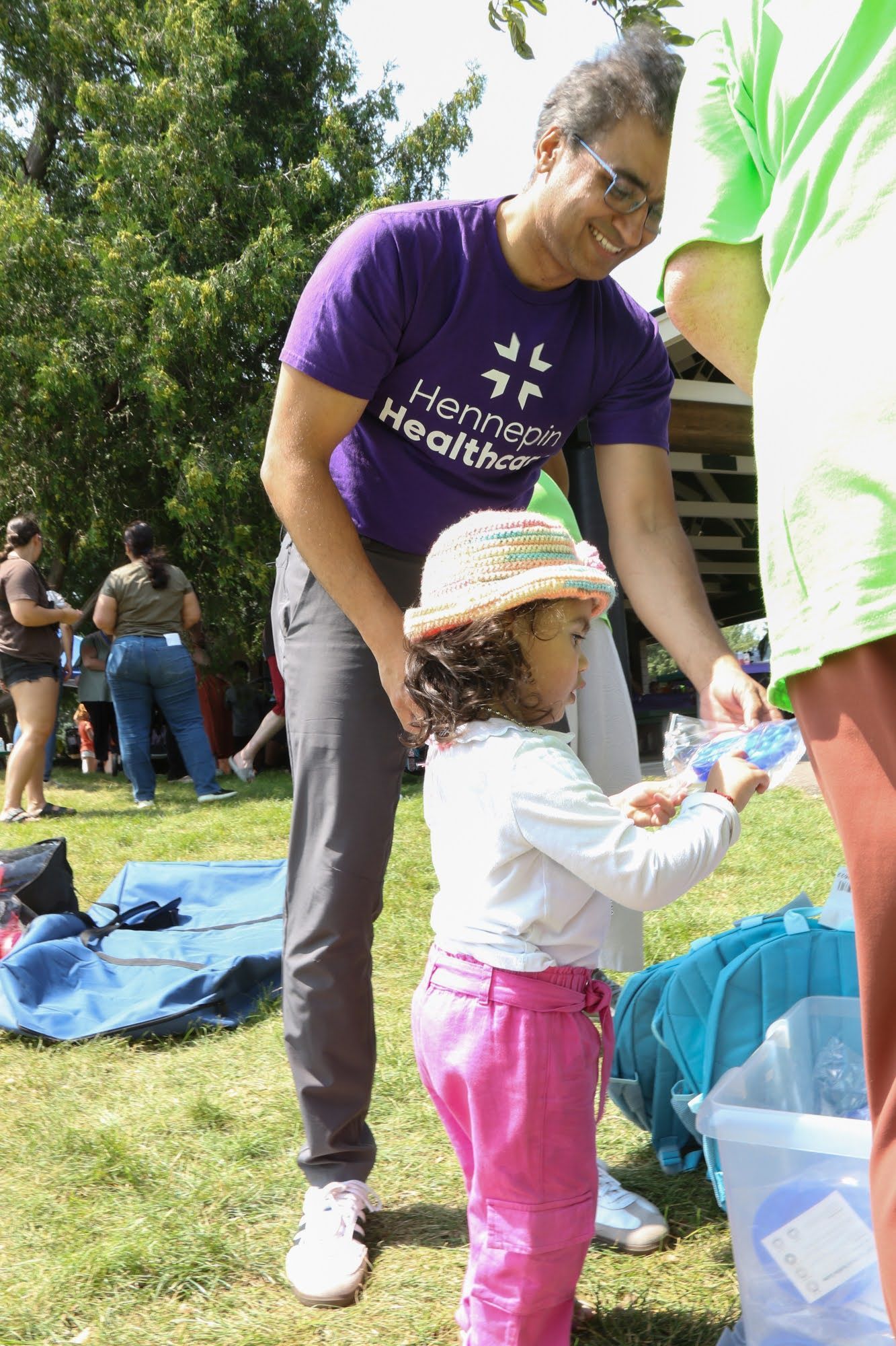 Man in purple shirt helps child in pink pants. Outdoors, sunny day.