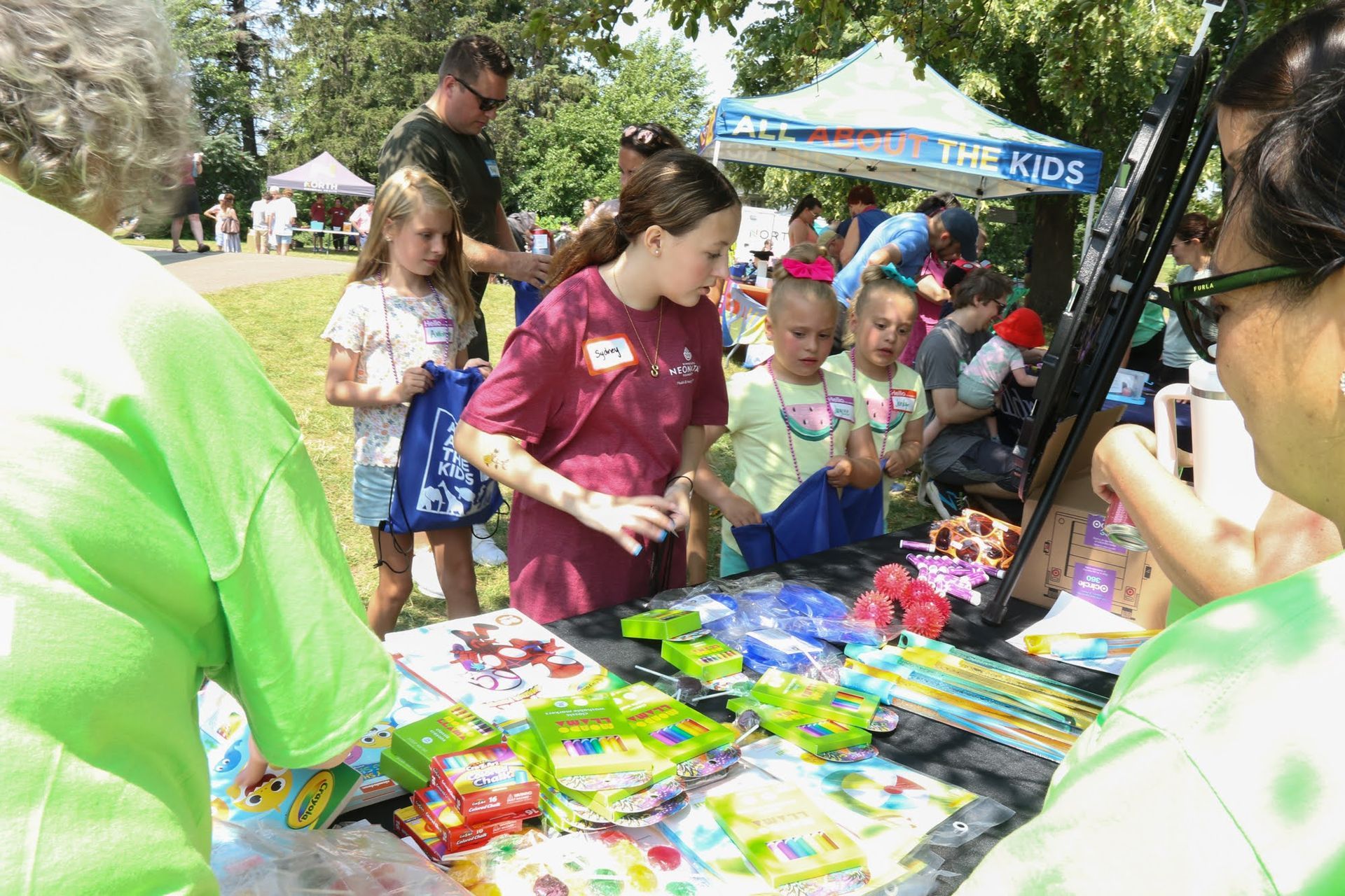 Children looking at items on a table at an outdoor event, people nearby, sunny day.