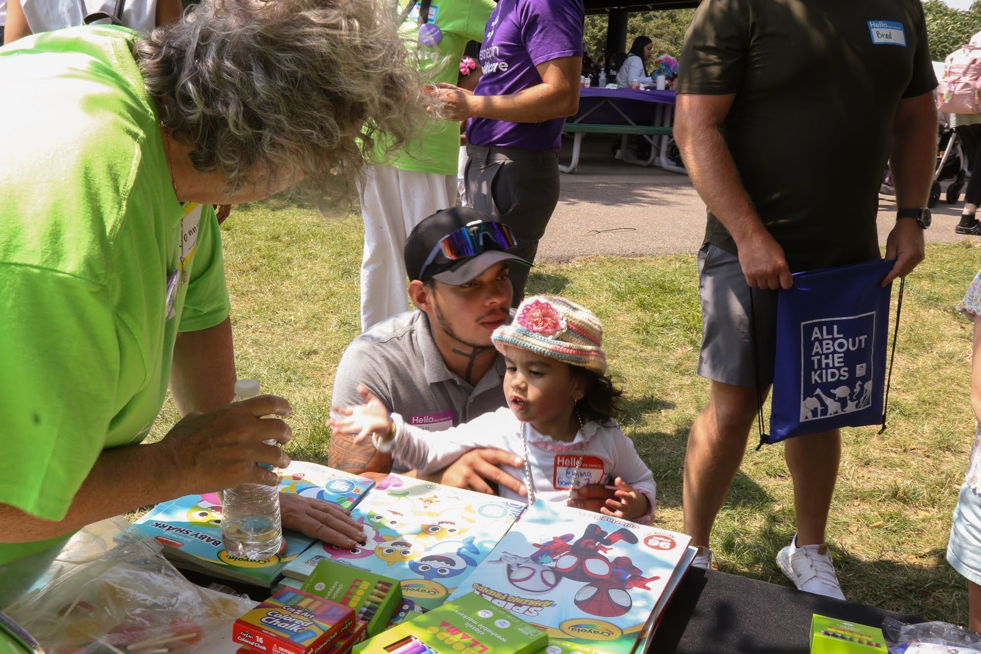 A child sits at a table with an adult; another adult leans over. Outdoor event with others.