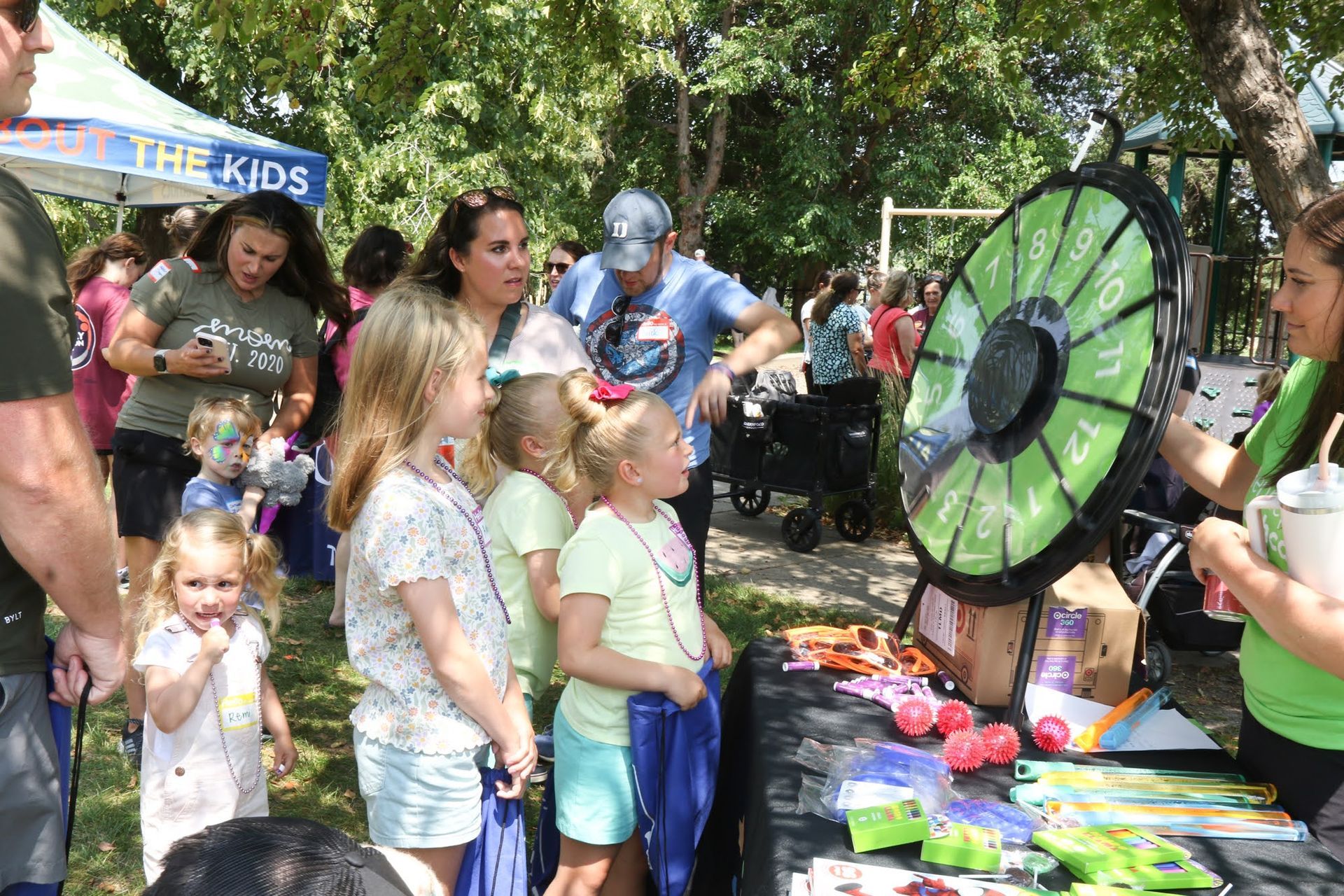 People at a fair booth with a prize wheel. Children watch as a woman spins it. Other people in the background.