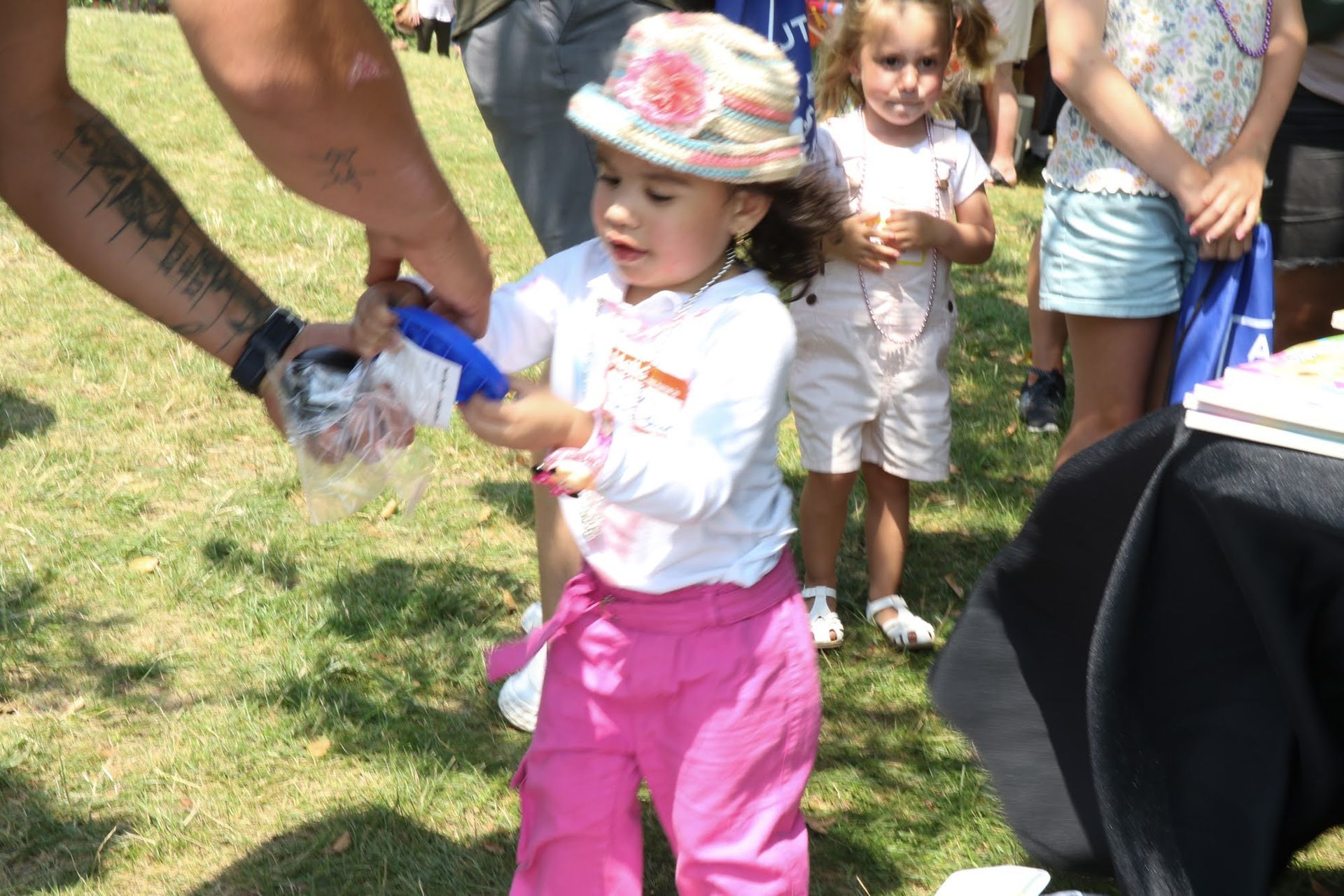 Child receiving an item from an adult at an outdoor event, wearing pink pants and a hat.