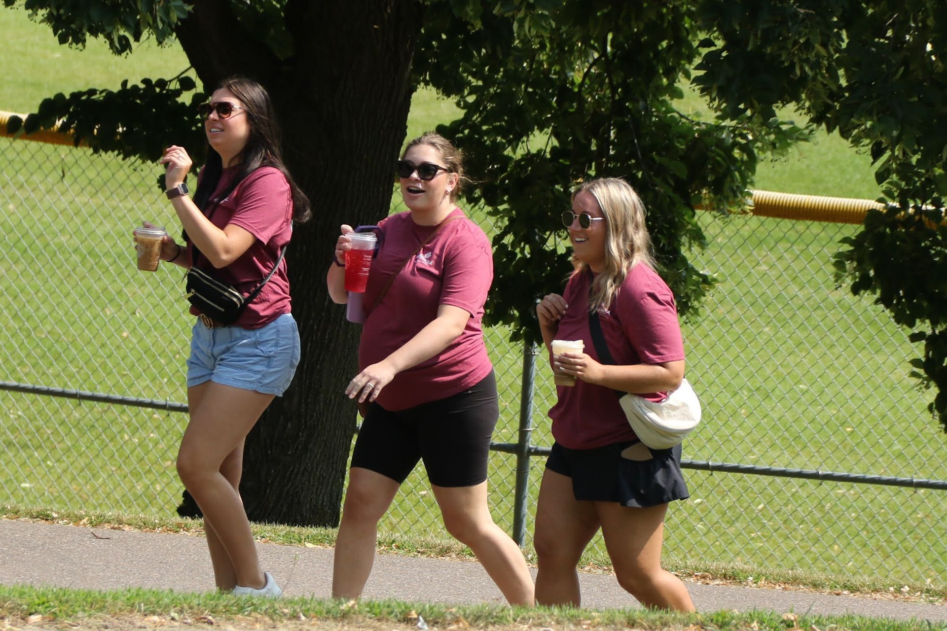 Three people in maroon shirts and sunglasses walking on a path in a park, each holding a beverage.