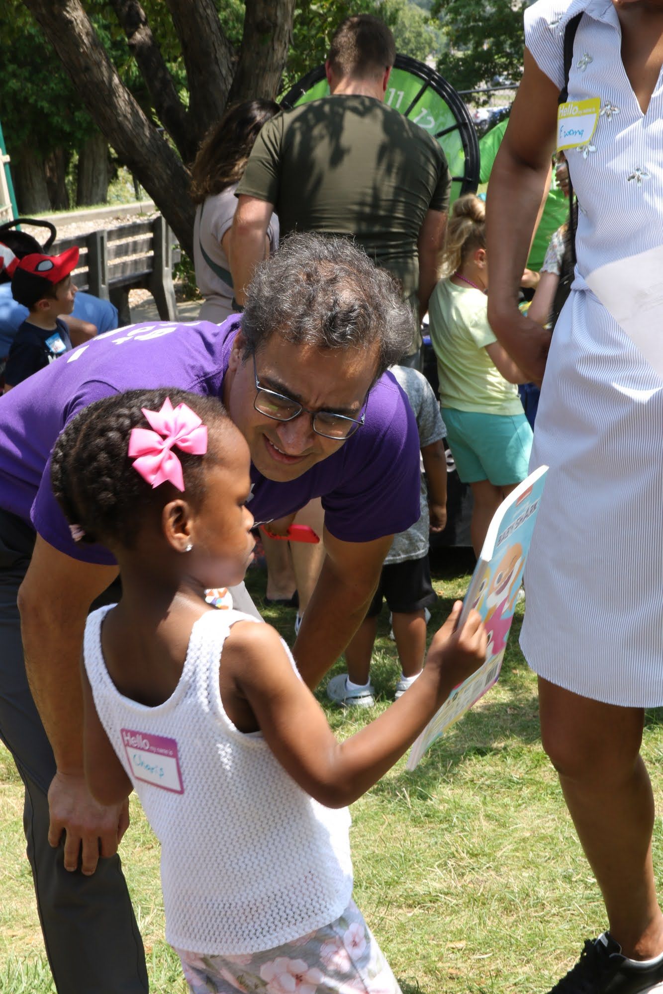 Man in purple shirt leans to look at a young girl holding a paper, outside. People, green background.