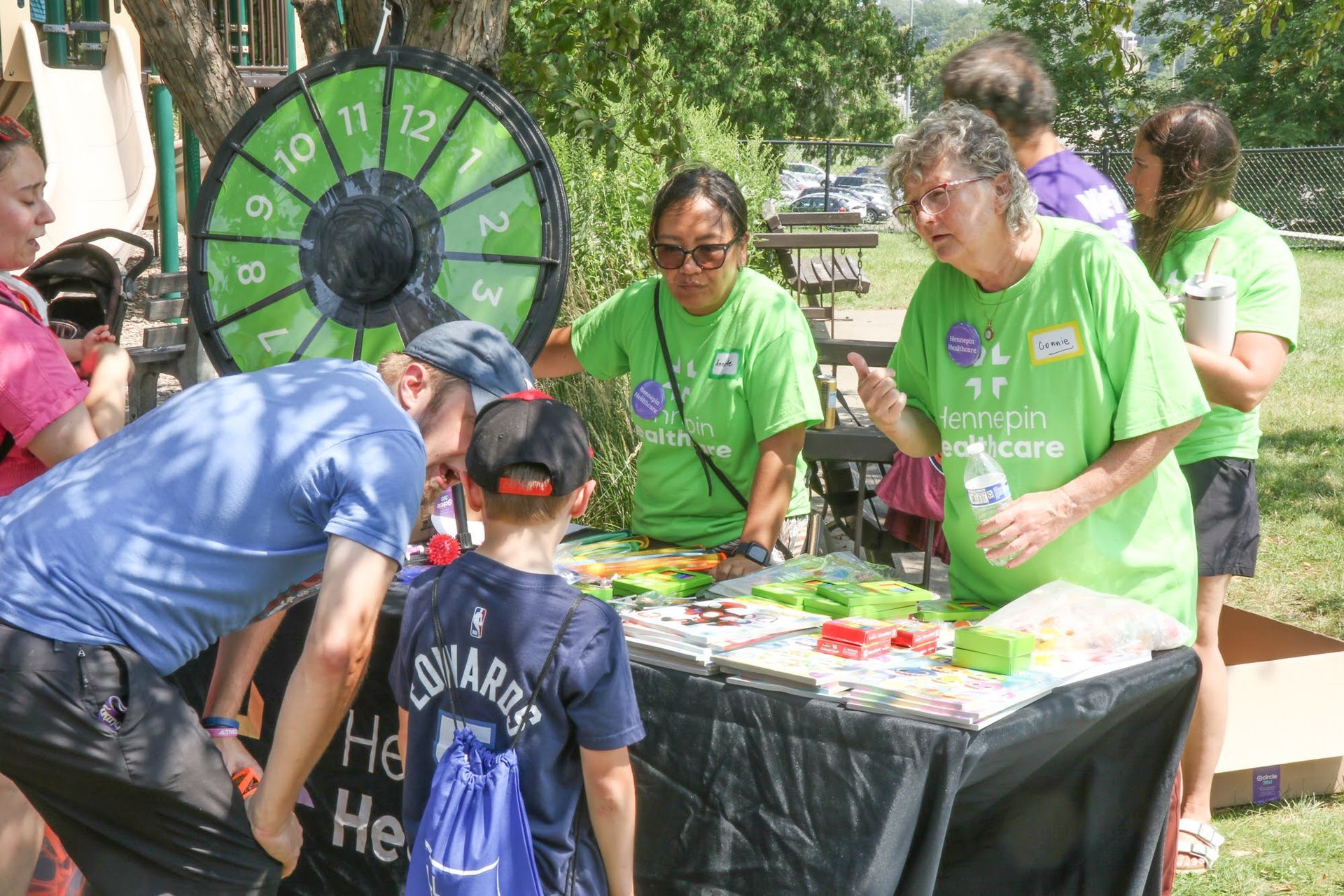 People at an outdoor table with a prize wheel. Two women in green shirts assist; others look at a board game.