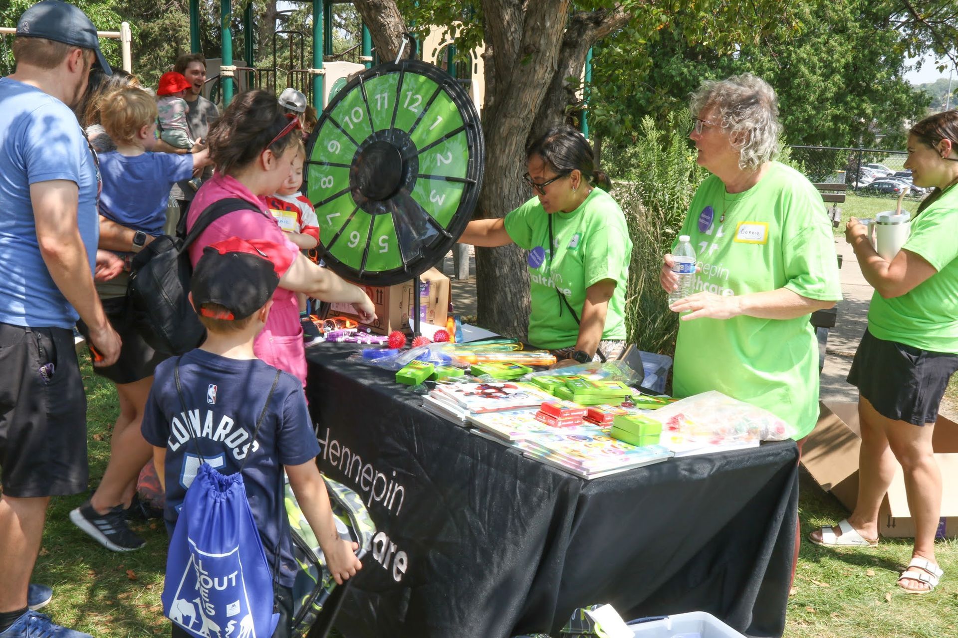 People at an outdoor event with a prize wheel and table, volunteers in green shirts, and toys.