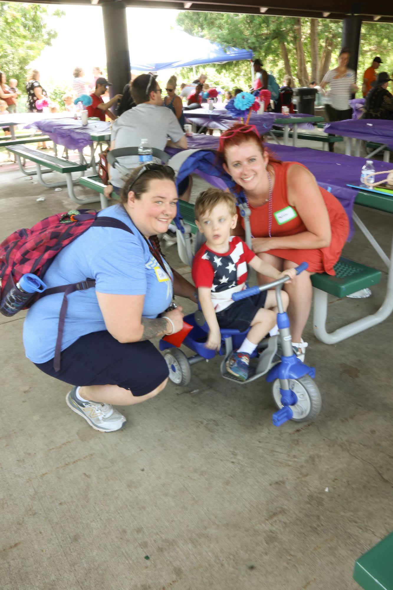 Two women and a child on a tricycle at a picnic. The child is smiling. People and tables are in the background.