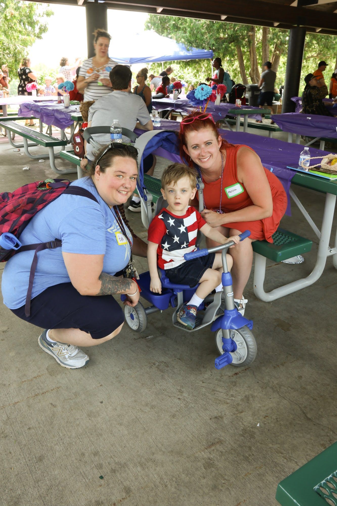 Two women and a child pose near a trike at a park with picnic tables.
