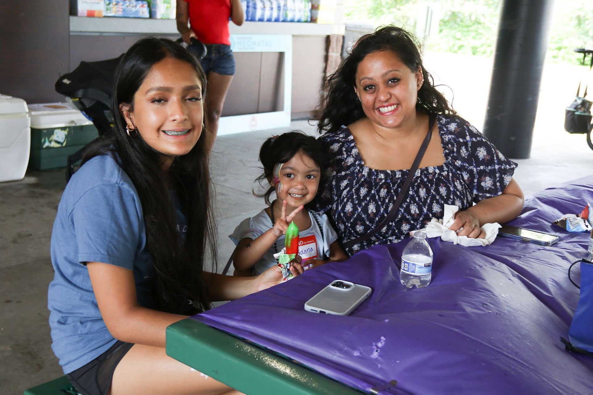 Three people smiling at a picnic table outdoors. One with braces. Purple tablecloth.