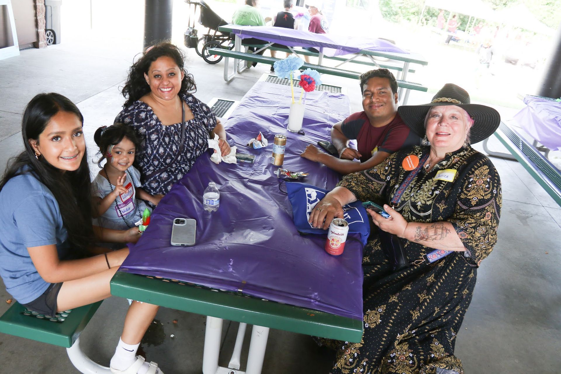 Group of people at picnic table under a shaded area, smiling.