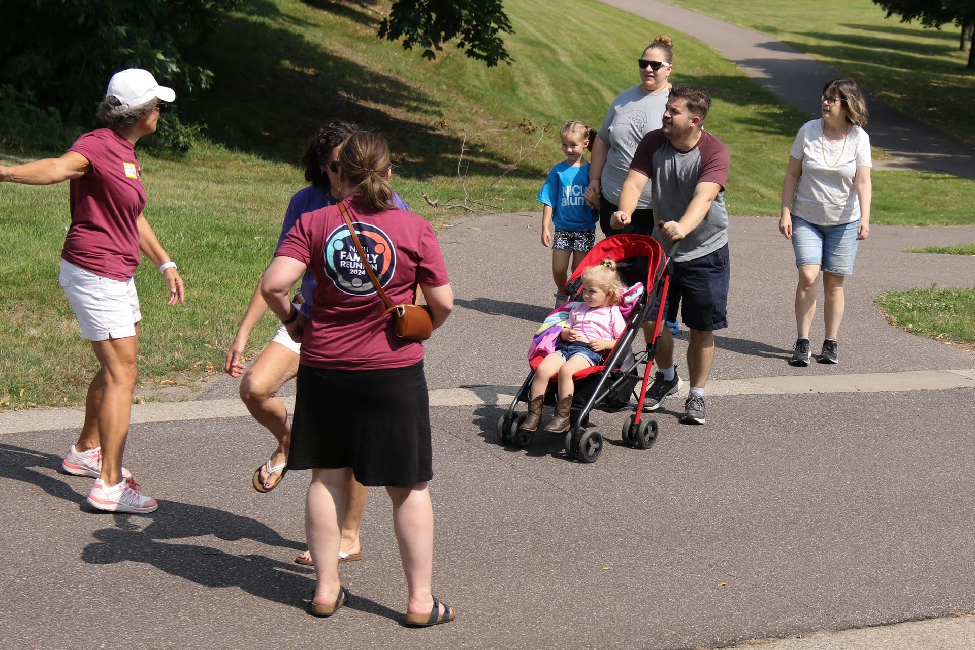 Group of people on a paved path in a park; one person with arms outstretched, another pushing a stroller.