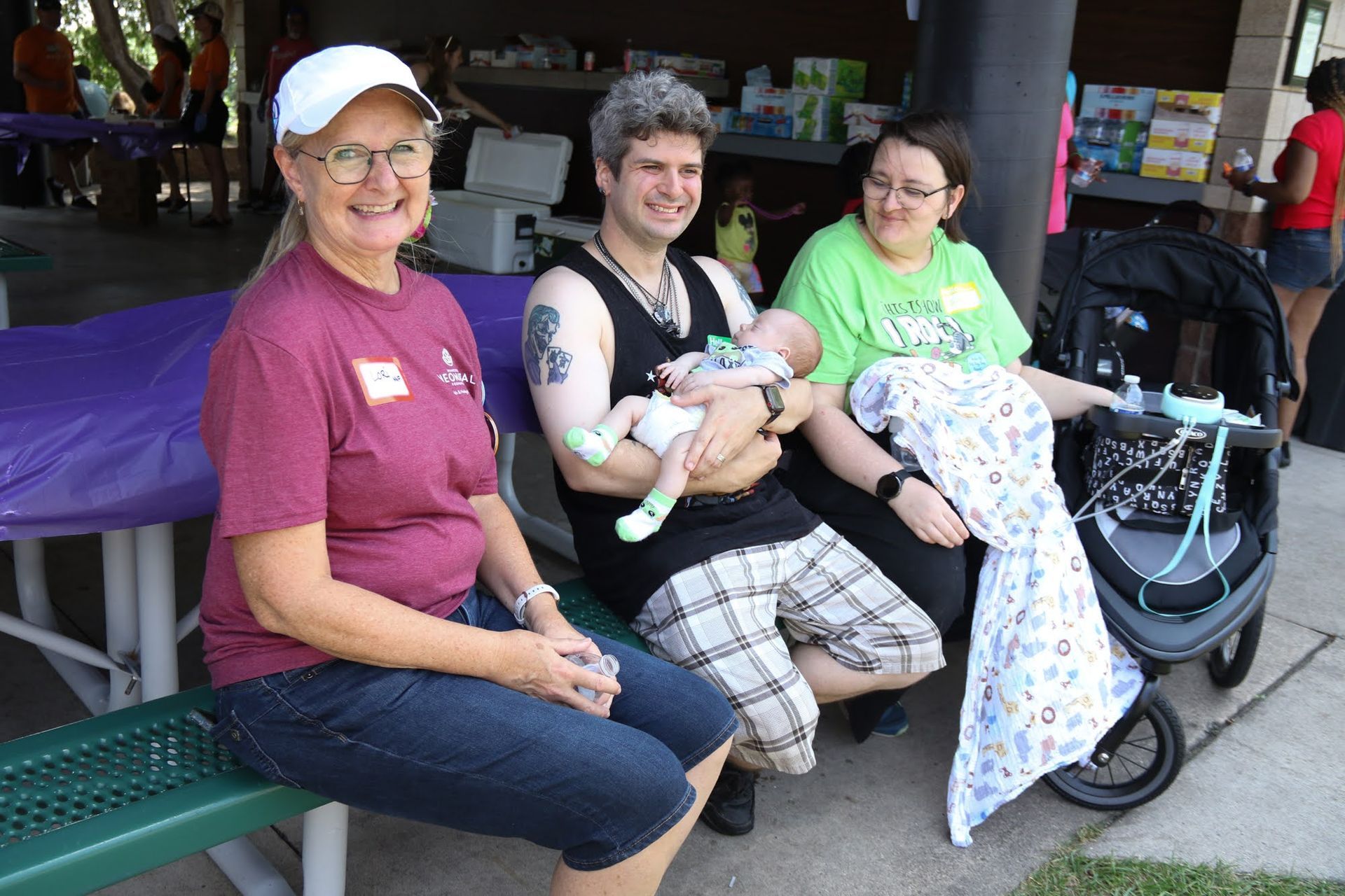 Three people and a baby outside, smiling. Man holds baby, woman in green sits with stroller.