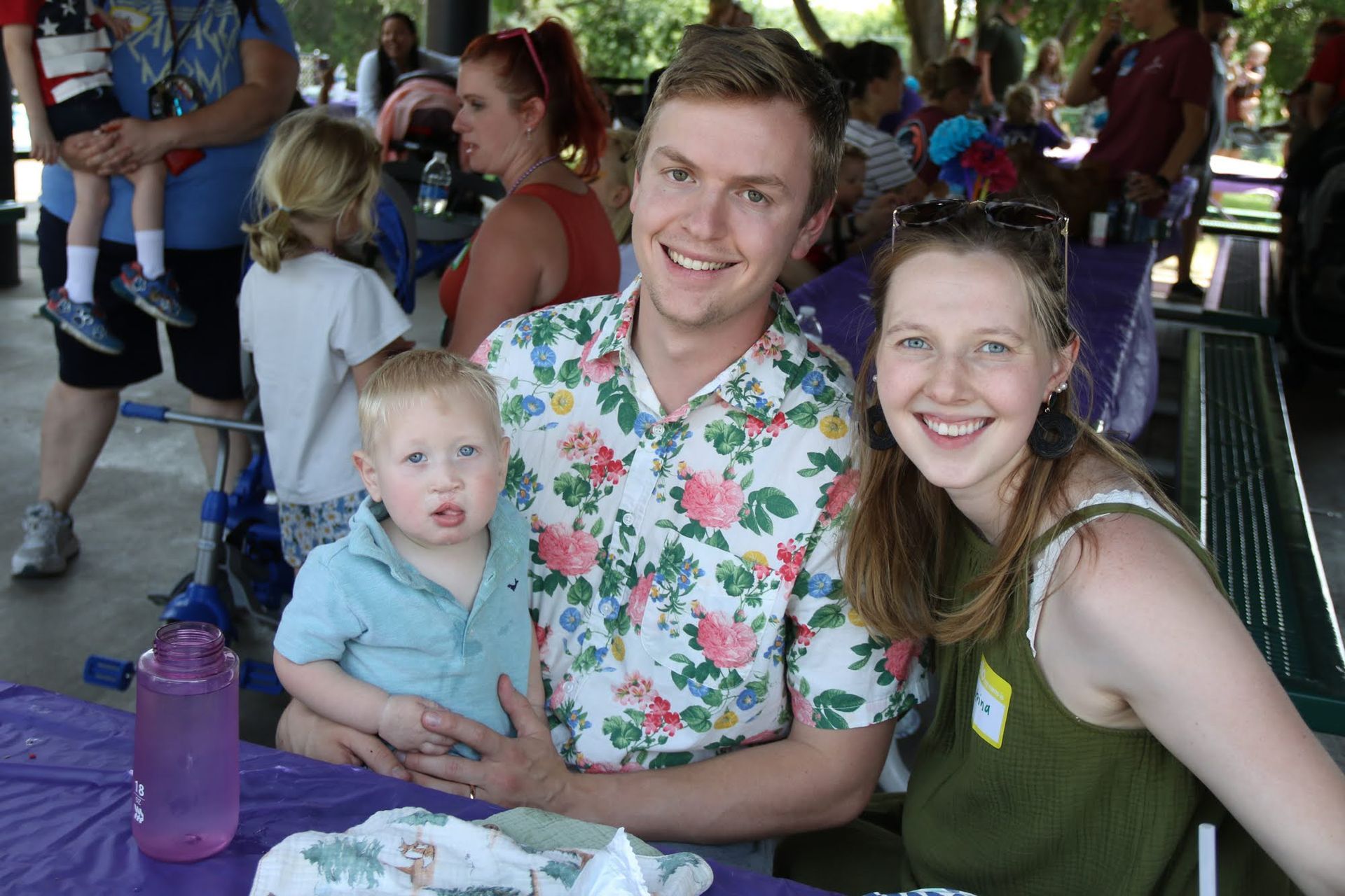Family at outdoor gathering, man in floral shirt holding toddler, woman smiles. Picnic table, other people.