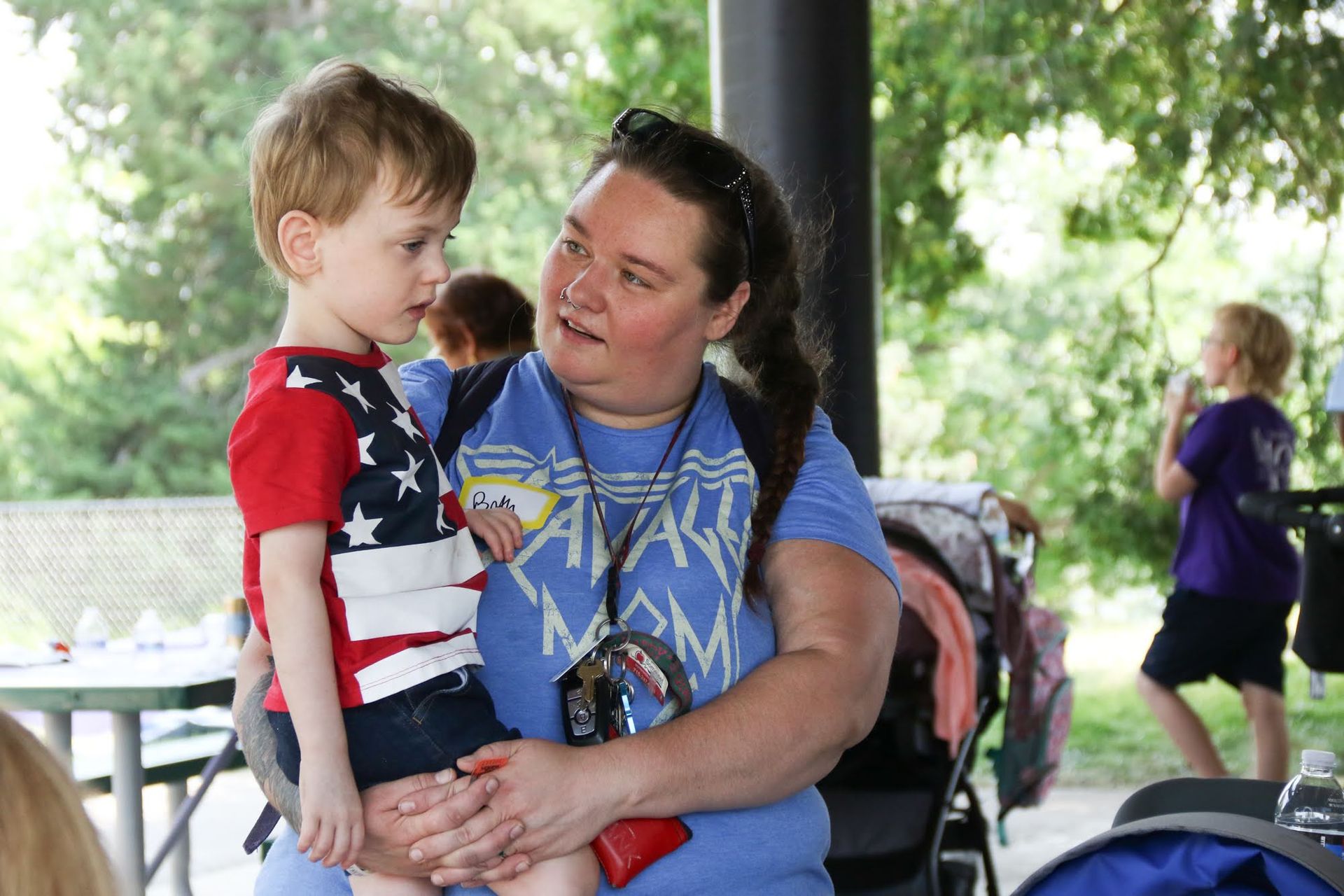 Woman holding a young child wearing an American flag shirt. They're outdoors, possibly at a park.