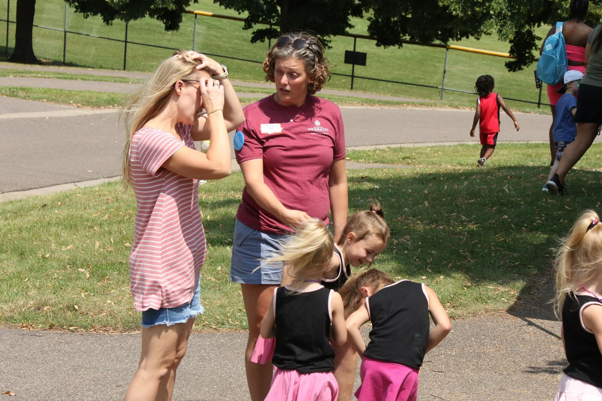 Two women talking to a group of children at a park. The woman on the left covers her eyes. Children play nearby.