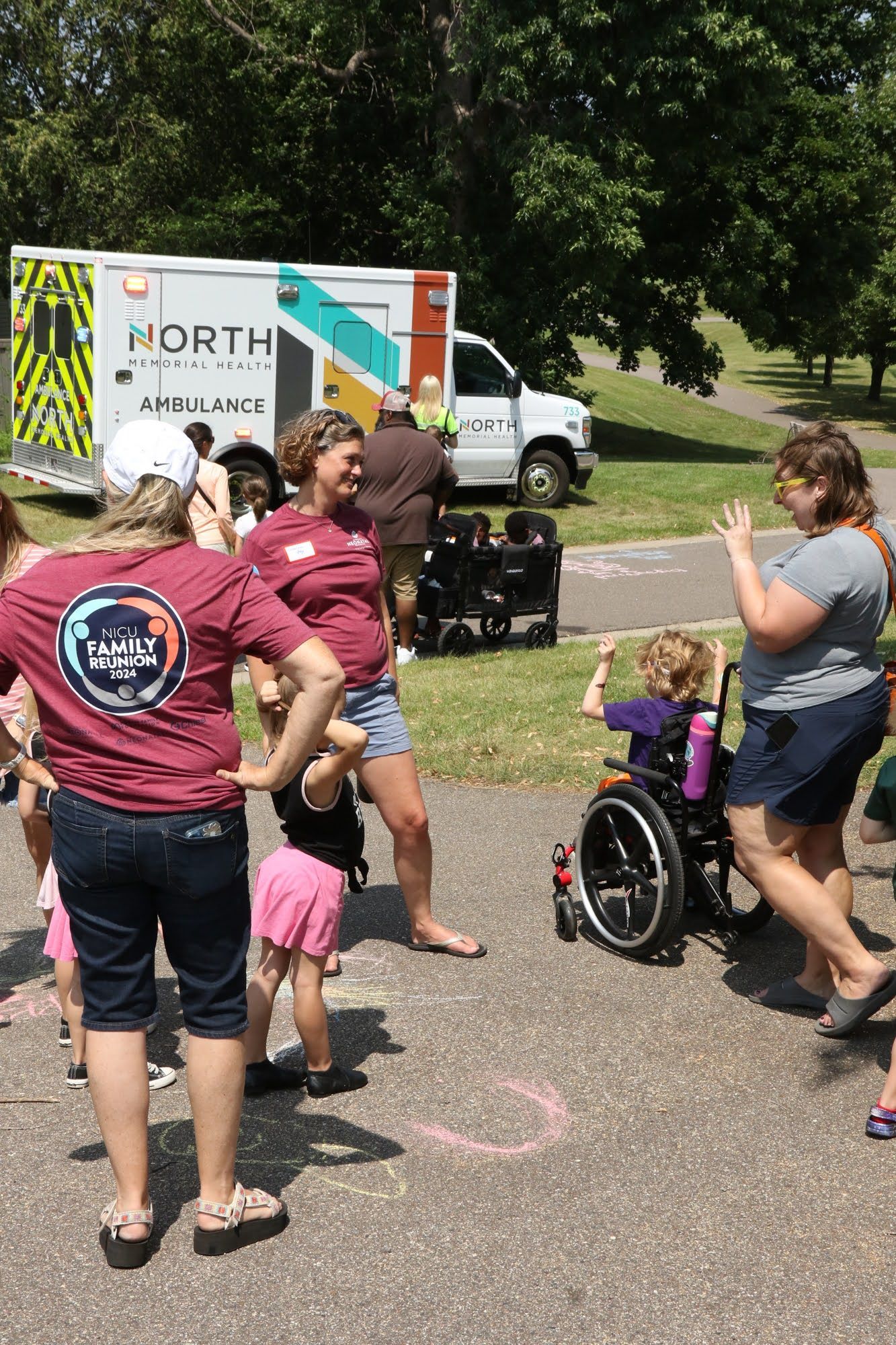 People gather near an ambulance and a child in a wheelchair. The child raises their arms, smiling. Outdoor setting.