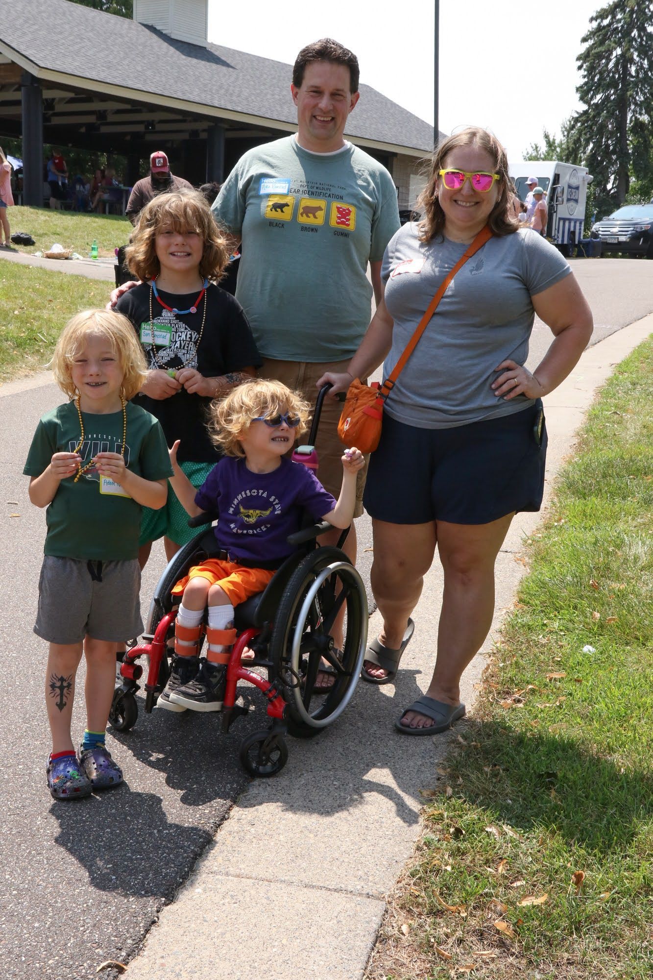 Family of five smiles near a path. One child uses a wheelchair. Outdoors, sunny day.