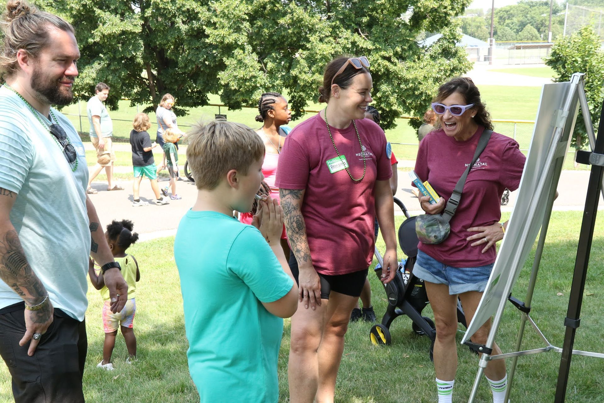 People at an outdoor event interact by a whiteboard. A boy smiles with his hands clasped as two women look on.