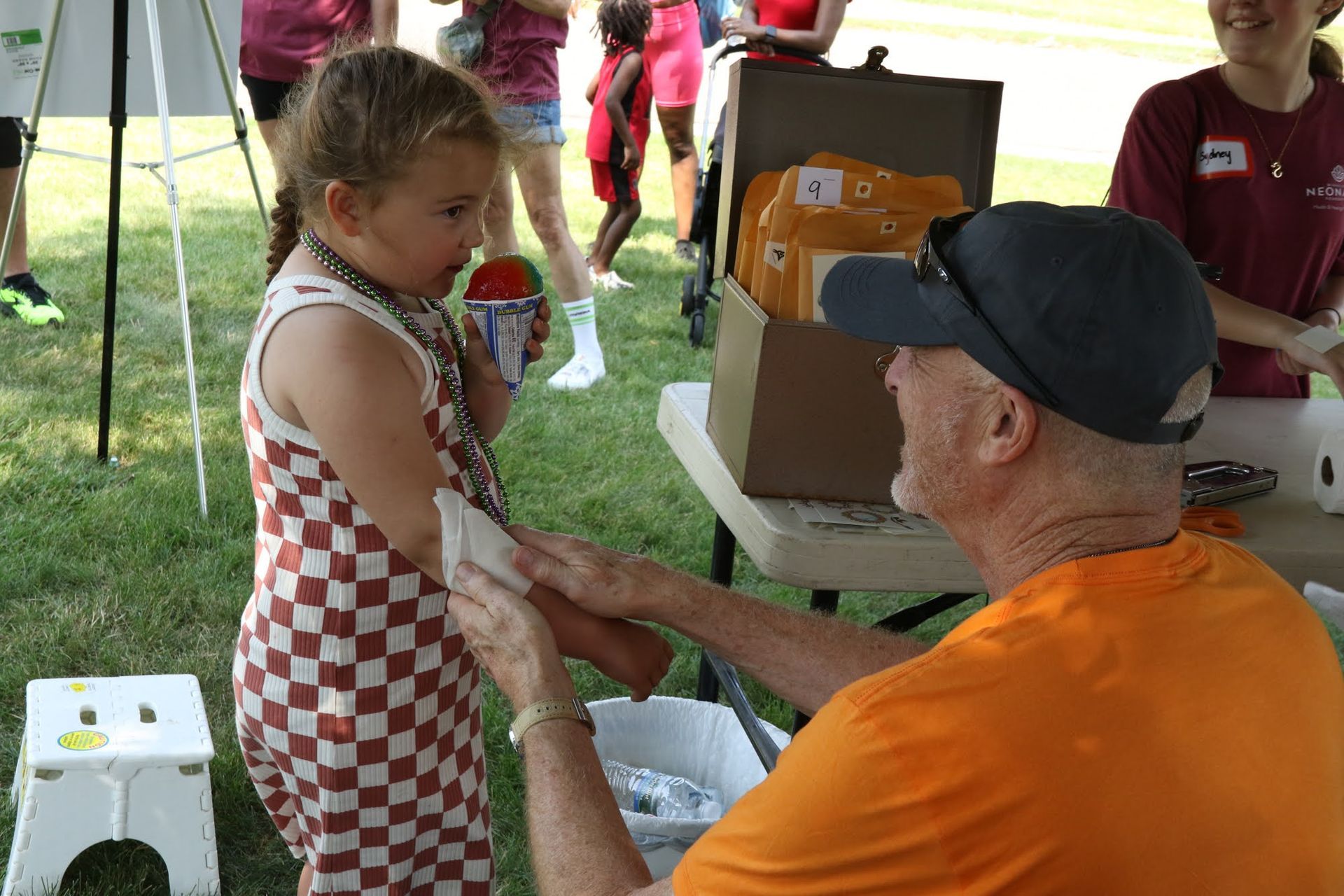 Girl with shaved ice gets a bandage from a man at an outdoor event.