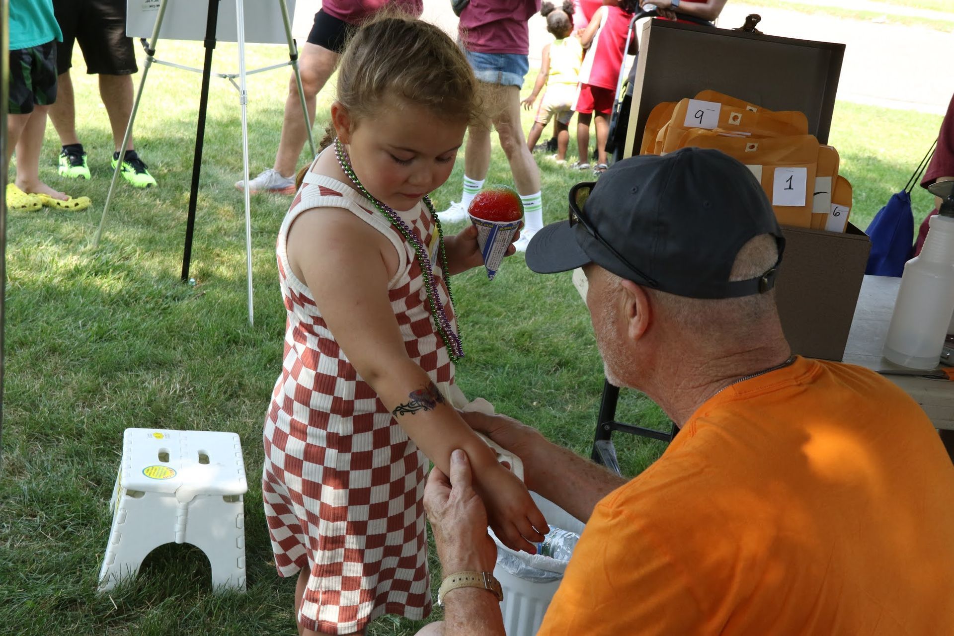 A girl in a checkered dress has her arm painted at an outdoor event, assisted by a man in an orange shirt.