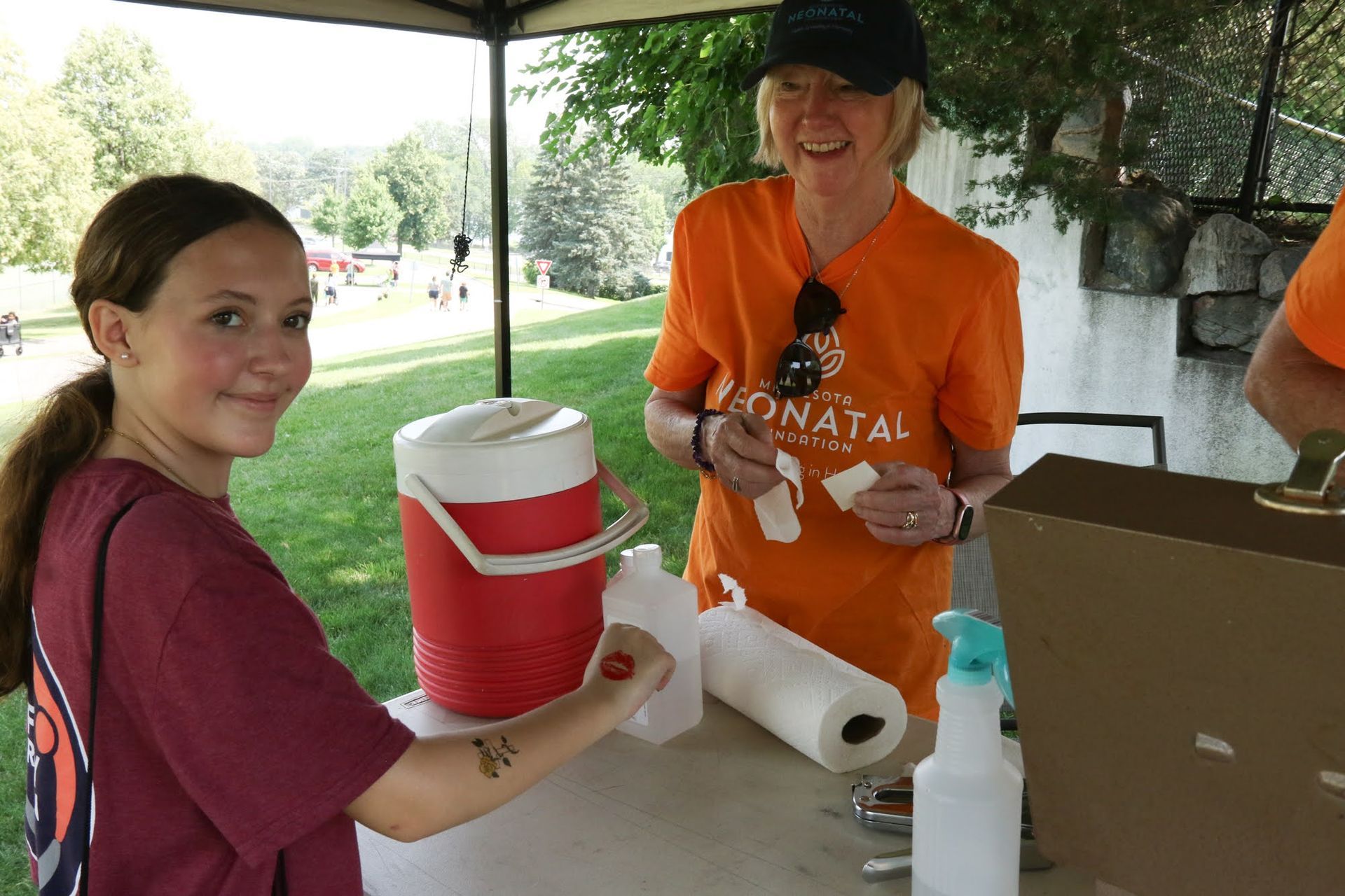 Two people at a table; one young person and one older person. They are smiling. Outdoor setting with cooler.