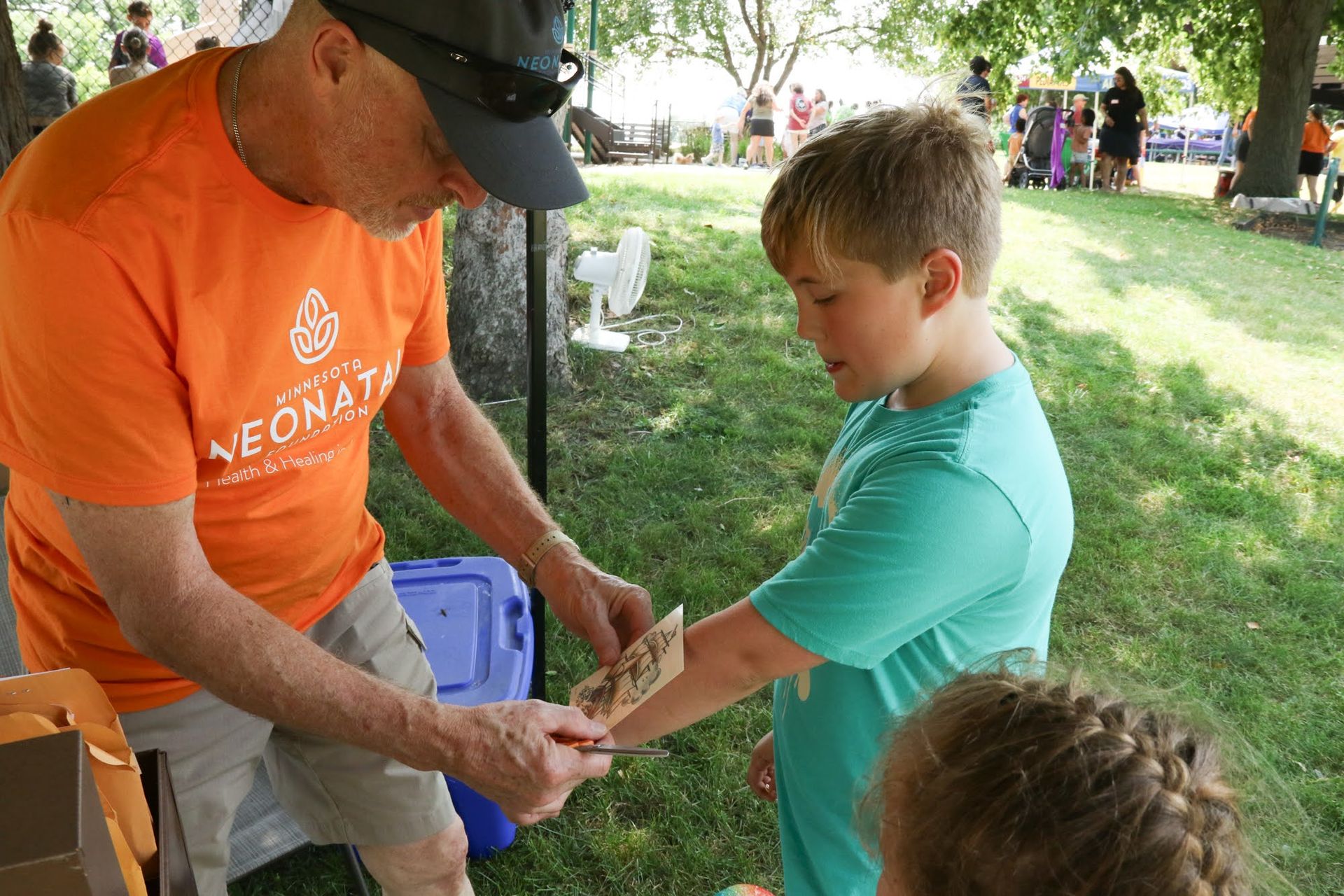 Man in orange shirt applies temporary tattoo to boy's arm outdoors.