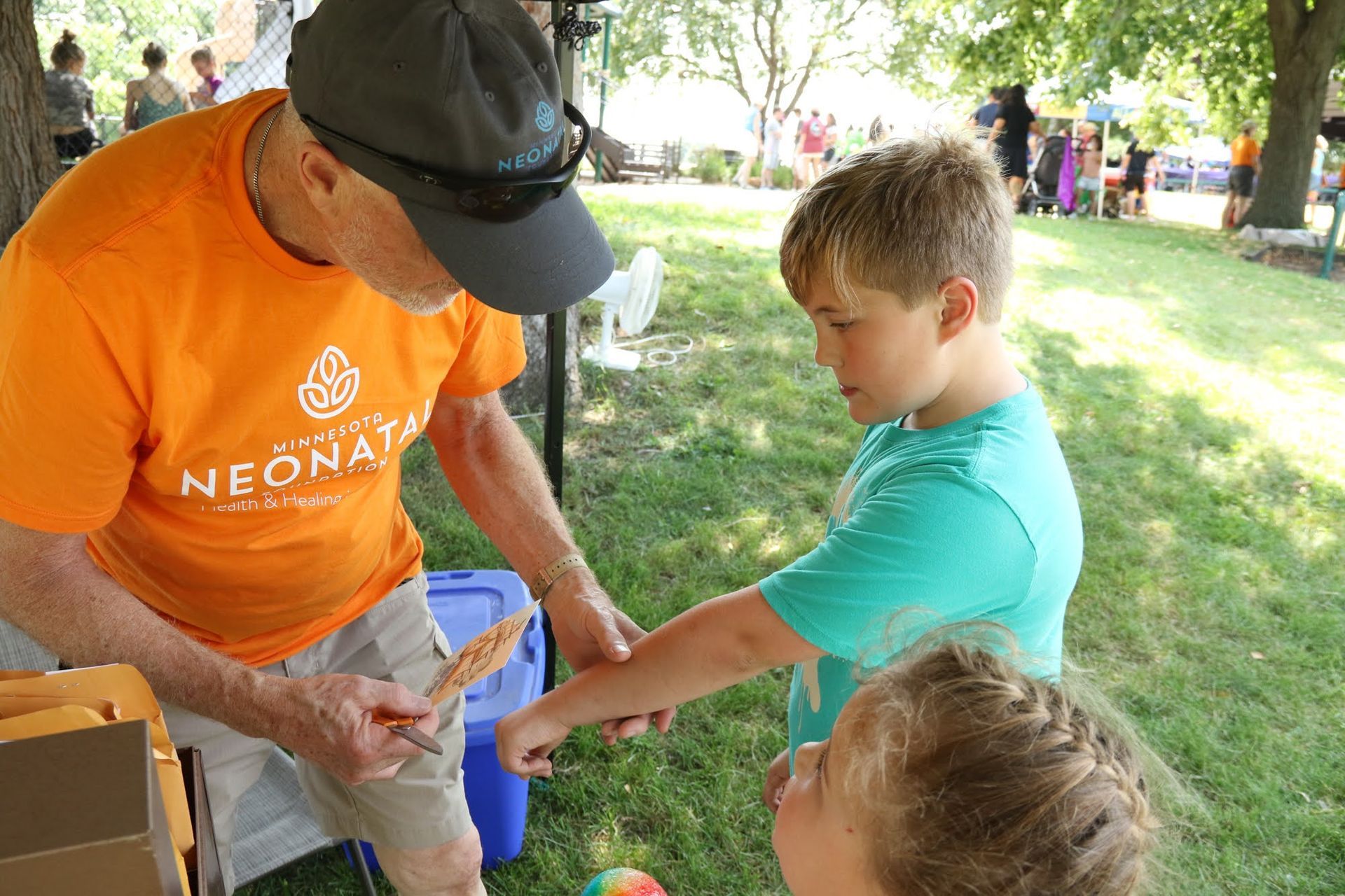 Man in orange shirt gives a popsicle to a child, another child watches nearby at an outdoor event.