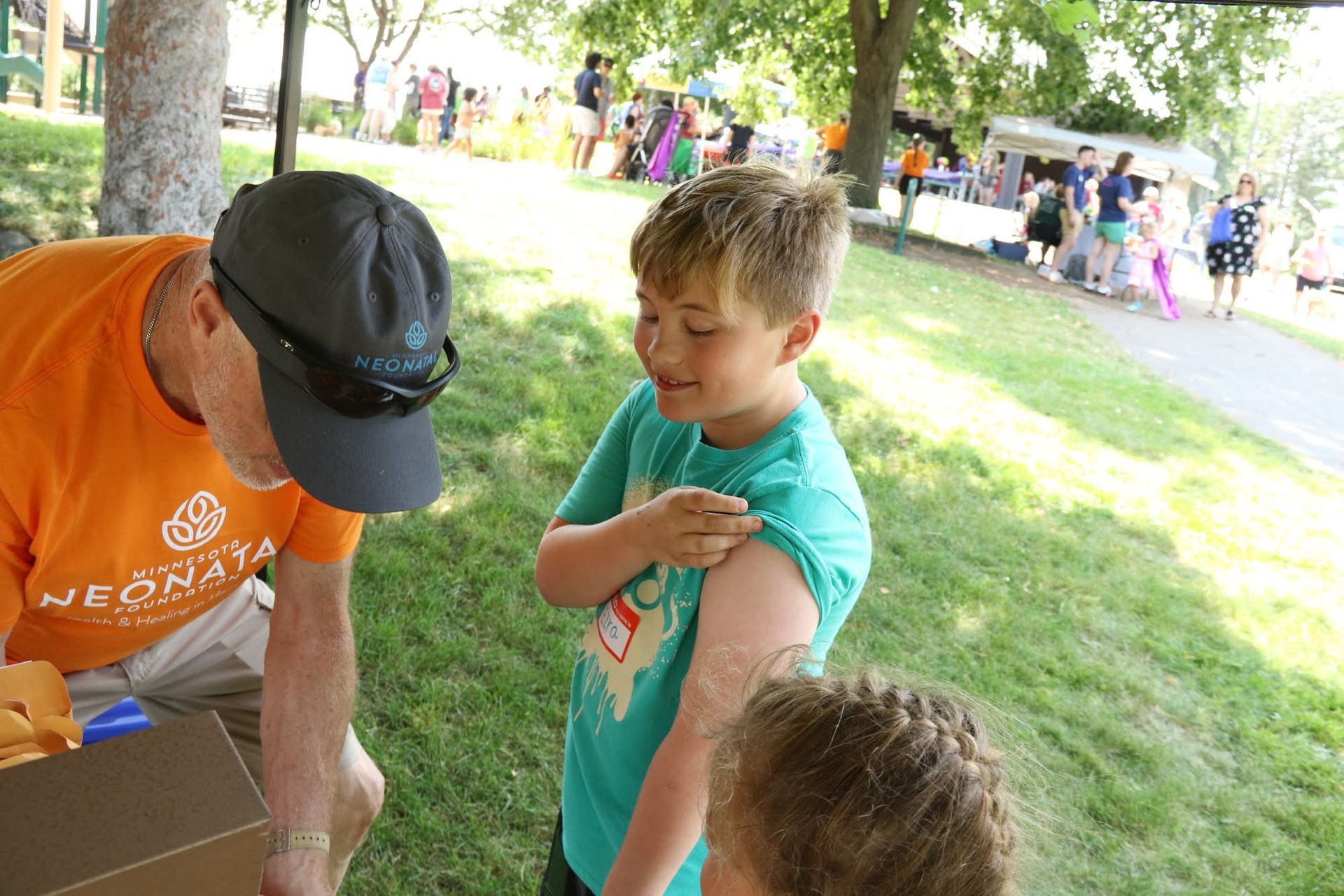 Man showing a temporary tattoo on a child's arm outdoors; child smiles; people in background.