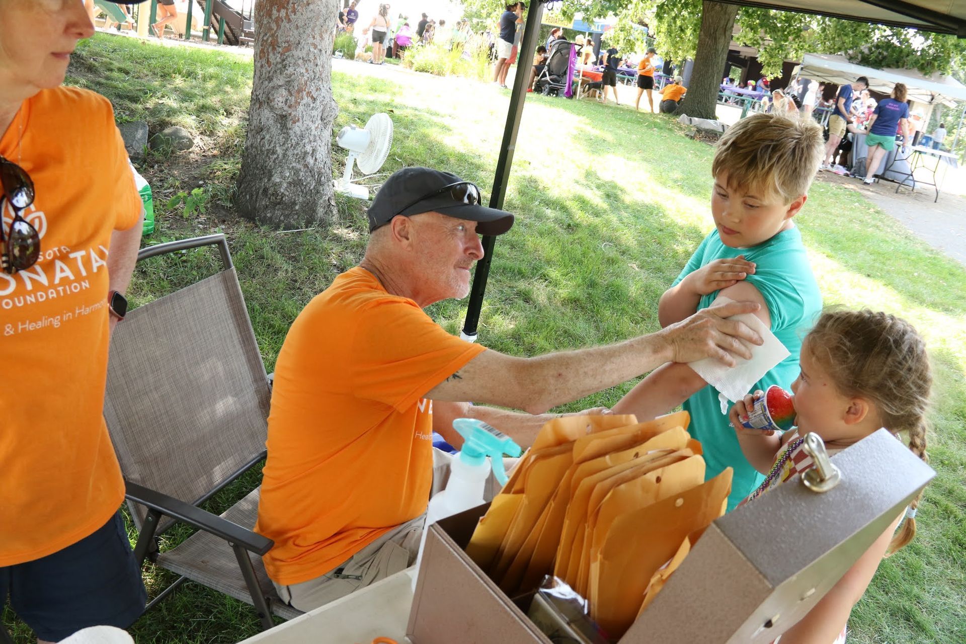 Man in orange shirt putting temporary tattoo on child's arm at outdoor event; another child watches.