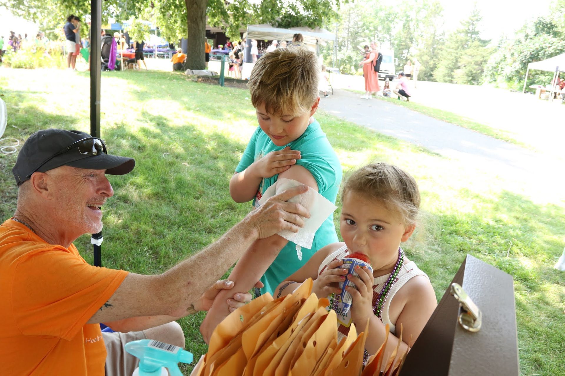 Man applying a temporary tattoo on a child's arm outdoors; another child drinks a beverage.