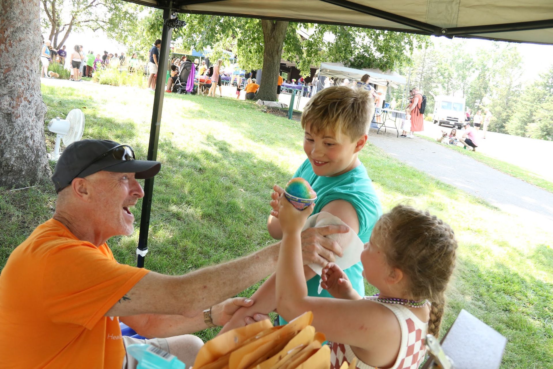 Man applying temporary tattoo to a child's arm at outdoor event. Boy and girl laugh excitedly under a canopy.