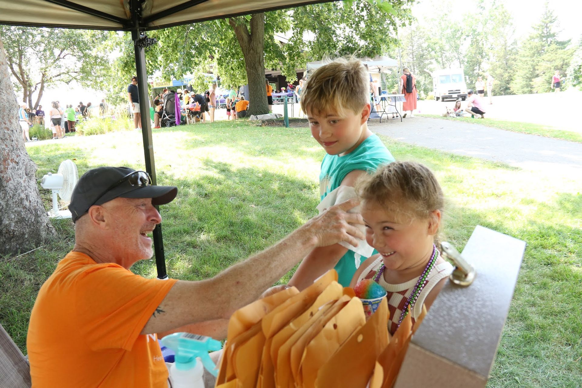 Man in orange shirt handing out items to two smiling children at a park.