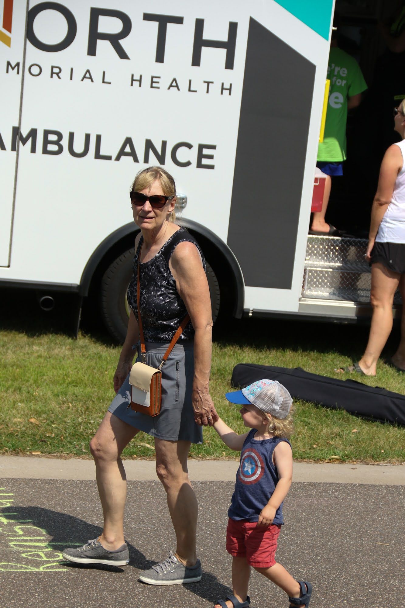 Woman in sunglasses holds hands with a young child. They walk past an ambulance.
